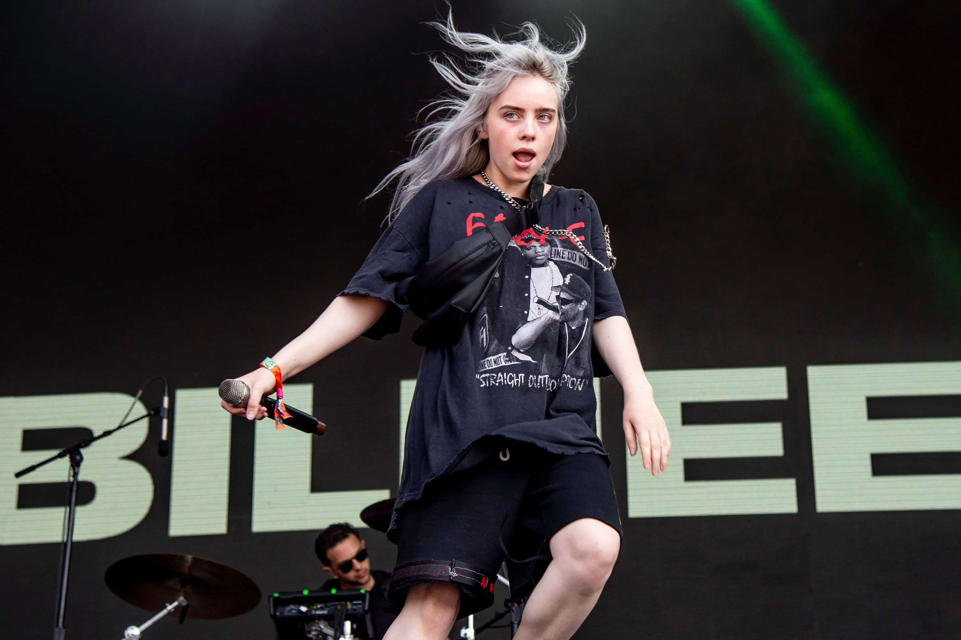 怪奇比莉 桌布 壁紙 — Billie Eilish 在明亮的舞台燈光下，以被風吹拂的銀色頭髮和黑色AC/DC T恤充滿活力地表演。