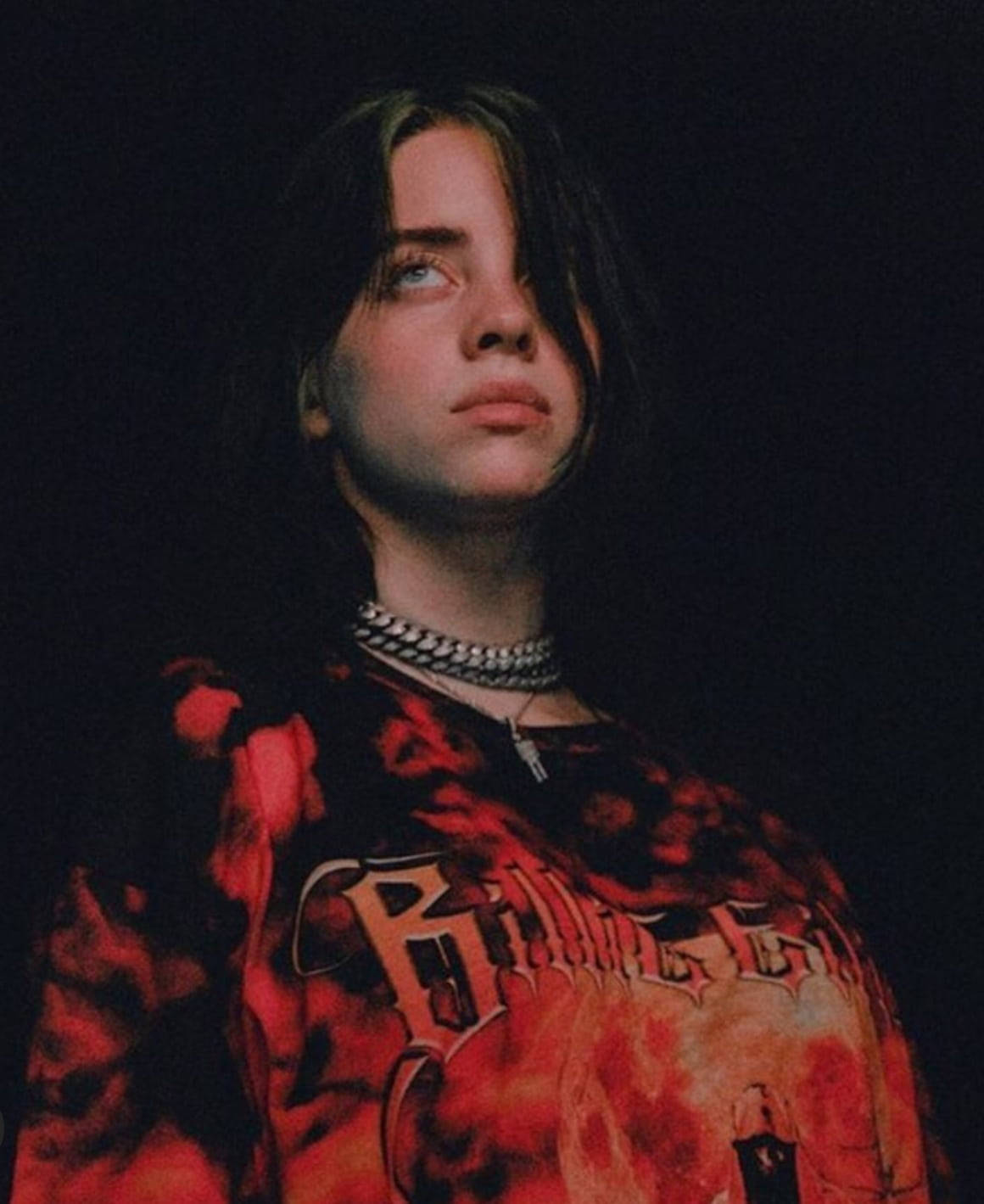 Billie Eilish 身穿紅色圖案T恤，戴著銀鏈，背景昏暗。 — 怪奇比莉 GIF 壁紙