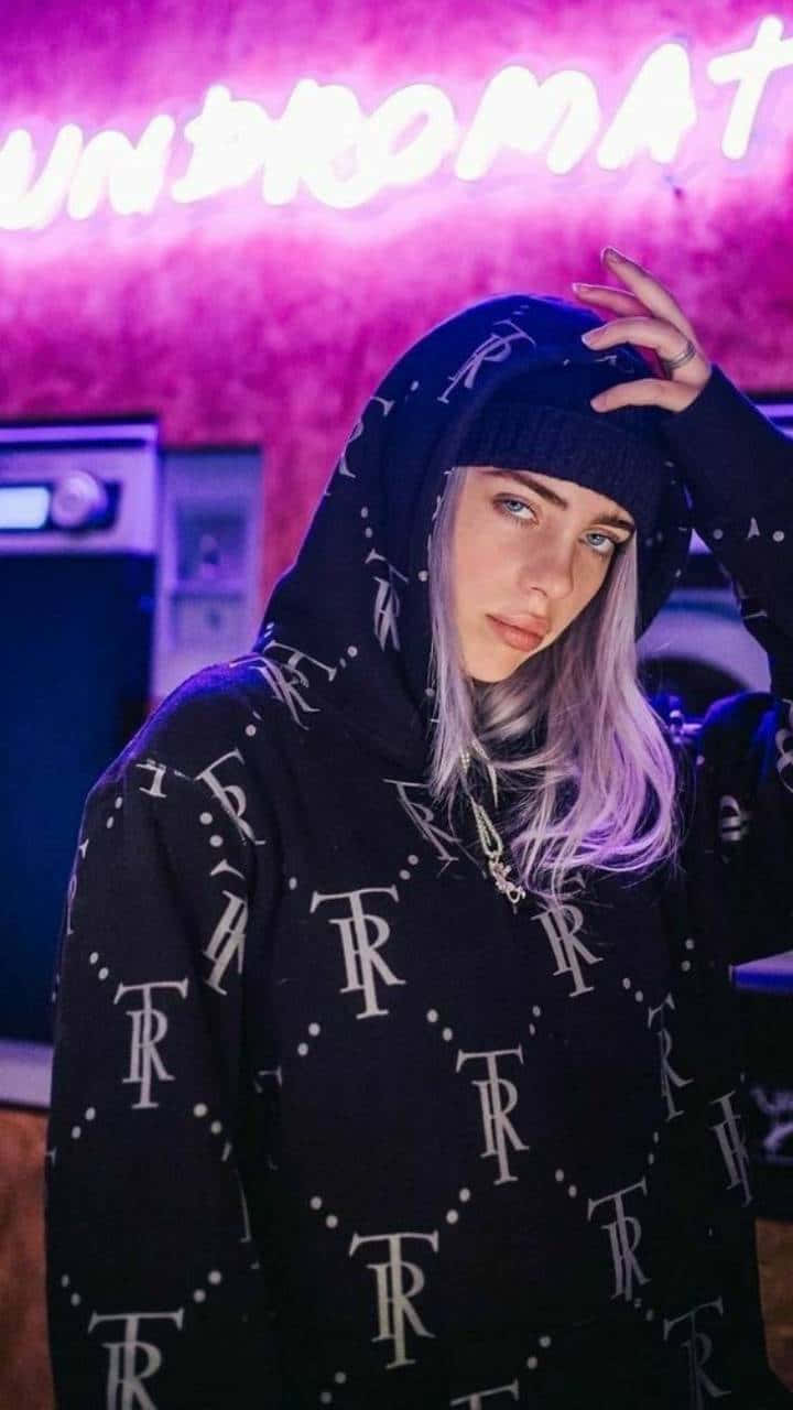 Billie Eilish Purple 桌布 壁紙 — Billie Eilish 在昏暗的房間裡，穿著帶有白色 TR 標誌的黑色連帽衫，在粉紅色霓虹燈下擺姿勢。