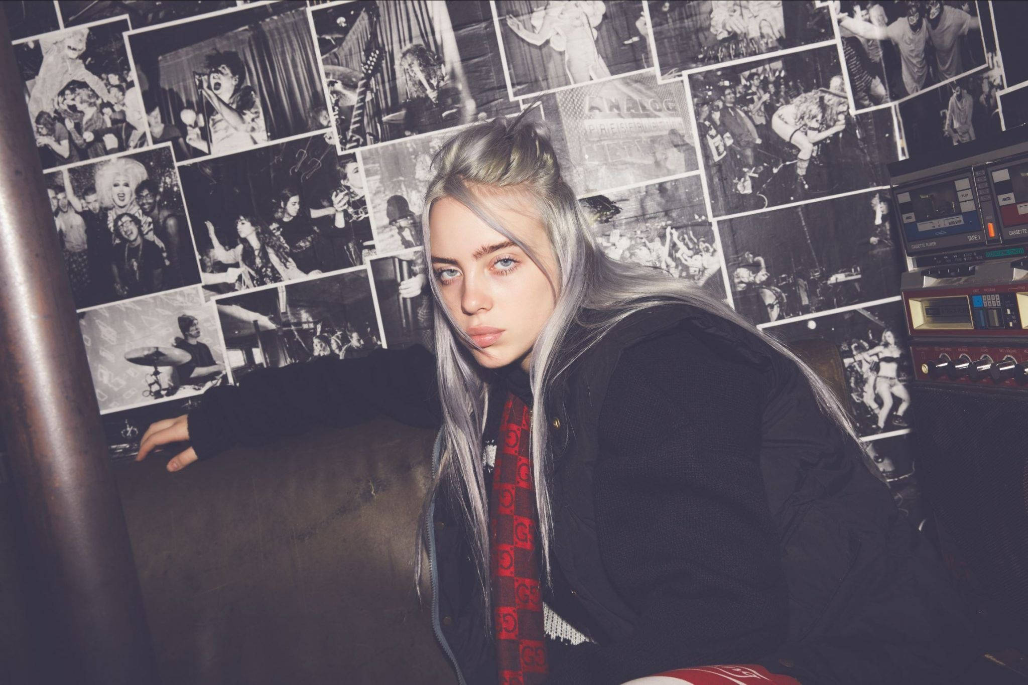 怪奇比莉 4K 桌布 壁紙 — Billie Eilish 在昏暗的房間裡，靠在一面鋪滿黑白演唱會照片的牆上。