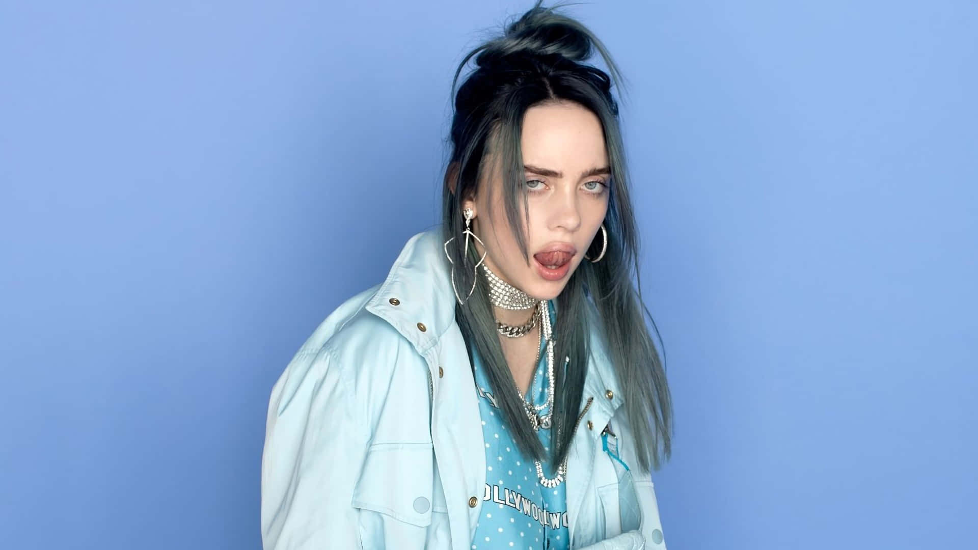Billie Eilish Laptop 桌布 壁紙 — Billie Eilish 在純藍色攝影棚背景前擺出俏皮表情，佩戴多層次珠寶和淺色牛仔外套。