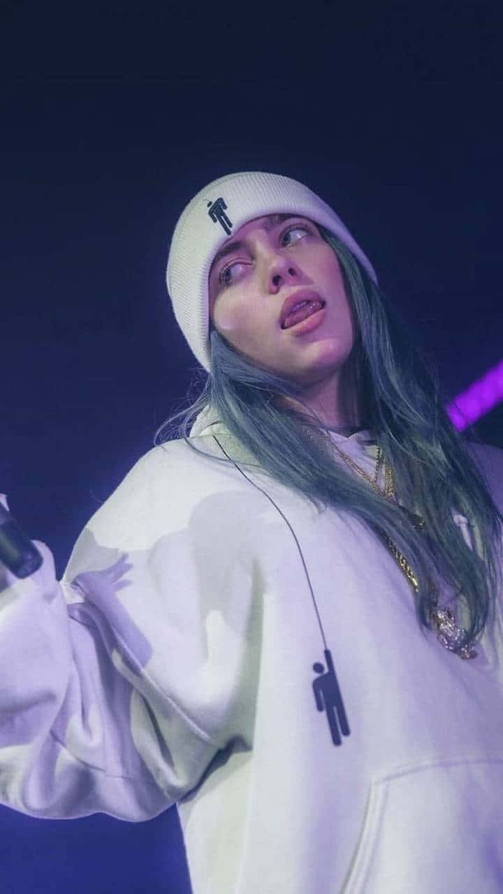 concert 桌布 壁紙 — Billie Eilish 在紫色舞台燈光下穿著白色連帽衫和毛帽在舞台上表演。