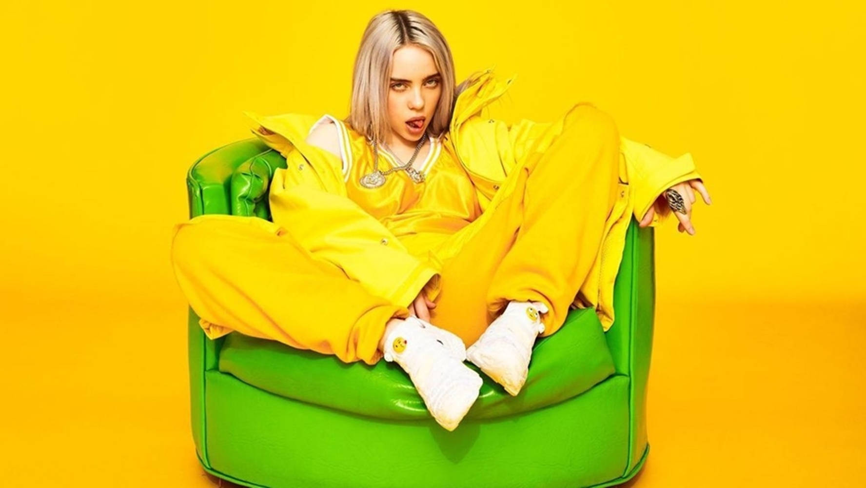 Billie Eilish 穿著黃色運動套裝，在黃色背景前坐在綠色椅子上。 — 怪奇比莉 GIF 壁紙