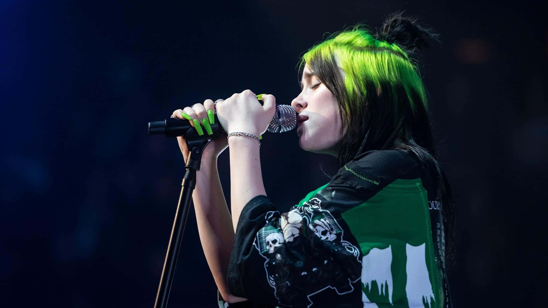 Billie Eilish Laptop 桌布 壁紙 — Billie Eilish 在黑暗的舞台燈光下，帶著充滿活力的綠色頭髮，熱情地對著麥克風表演。