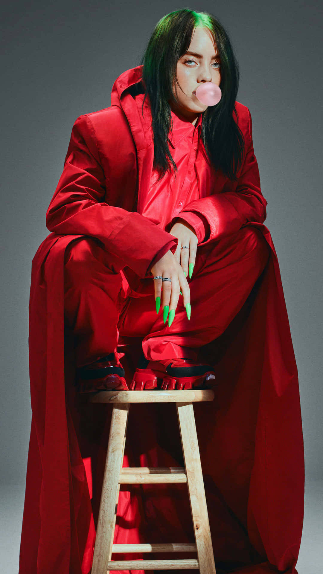 Billie Eilish 2021 桌布 壁紙 — Billie Eilish 坐在木凳上，身穿大膽的紅色服裝，在灰色工作室背景下吹出粉紅色泡泡糖泡泡。