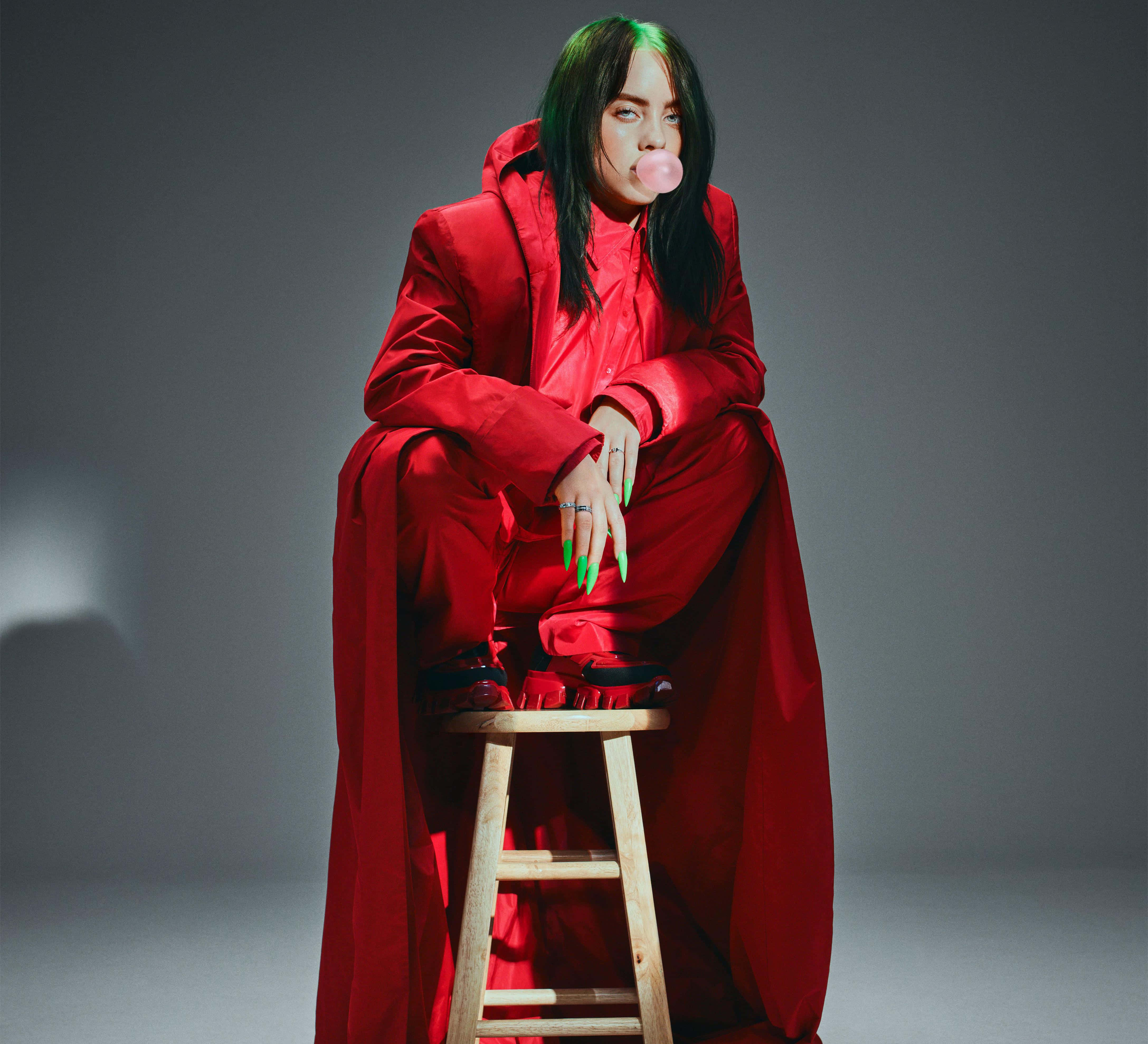 怪奇比莉 桌布 壁紙 — Billie Eilish 坐在木凳上，身穿大膽的紅色服裝，在灰色攝影棚背景下吹出粉紅色泡泡糖泡泡。