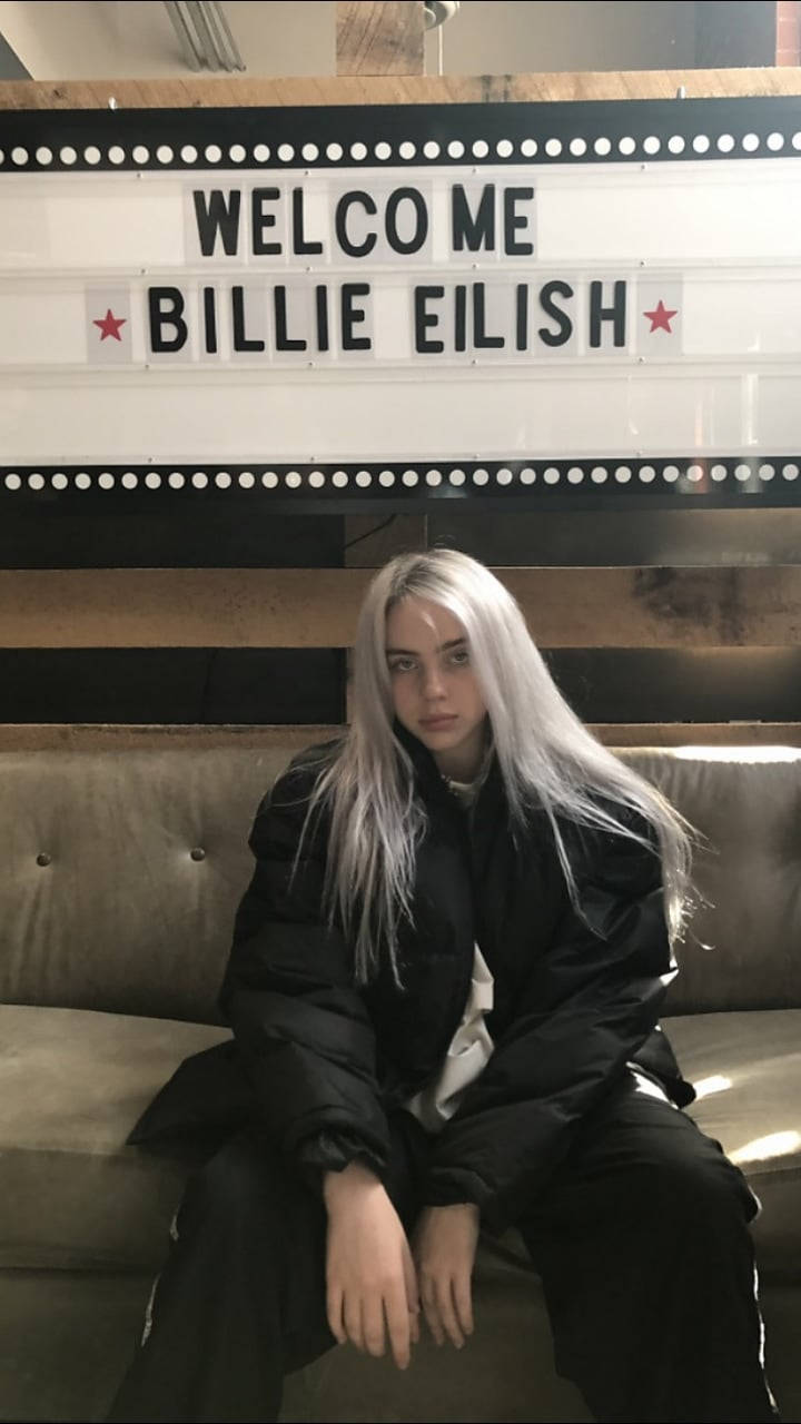 怪奇比莉 桌布 壁紙 — Billie Eilish 平靜地坐在米色沙發上，下方有一個發光的招牌，上面寫著 'WELCOME ME BILLIE EILISH' 並有紅色星星。
