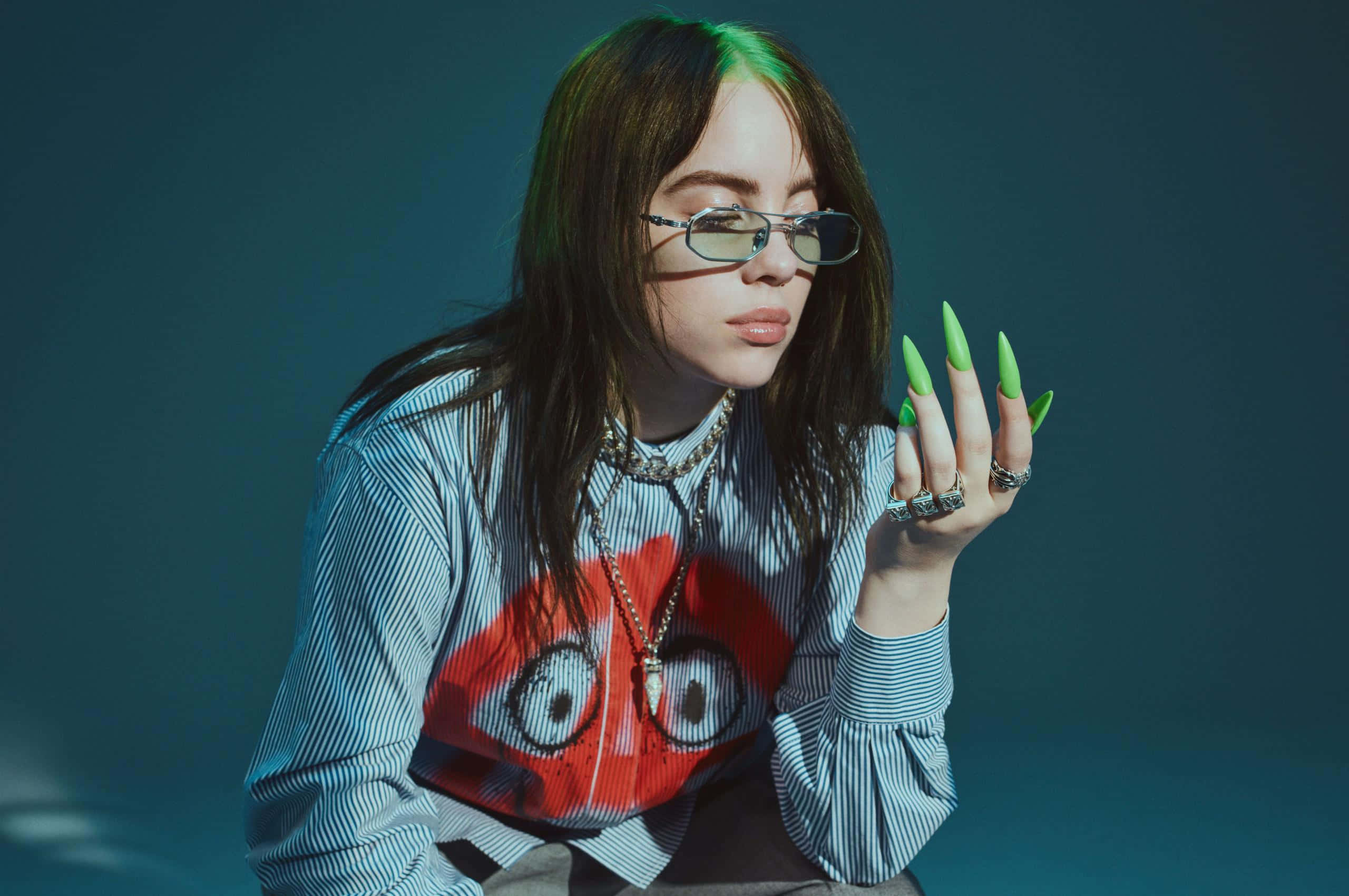 Billie Eilish Laptop 桌布 壁紙 — Billie Eilish 戴著眼鏡，留著綠色指甲，身穿帶有紅色眼睛圖案的條紋襯衫，在憂鬱的青色背景前擺姿勢。