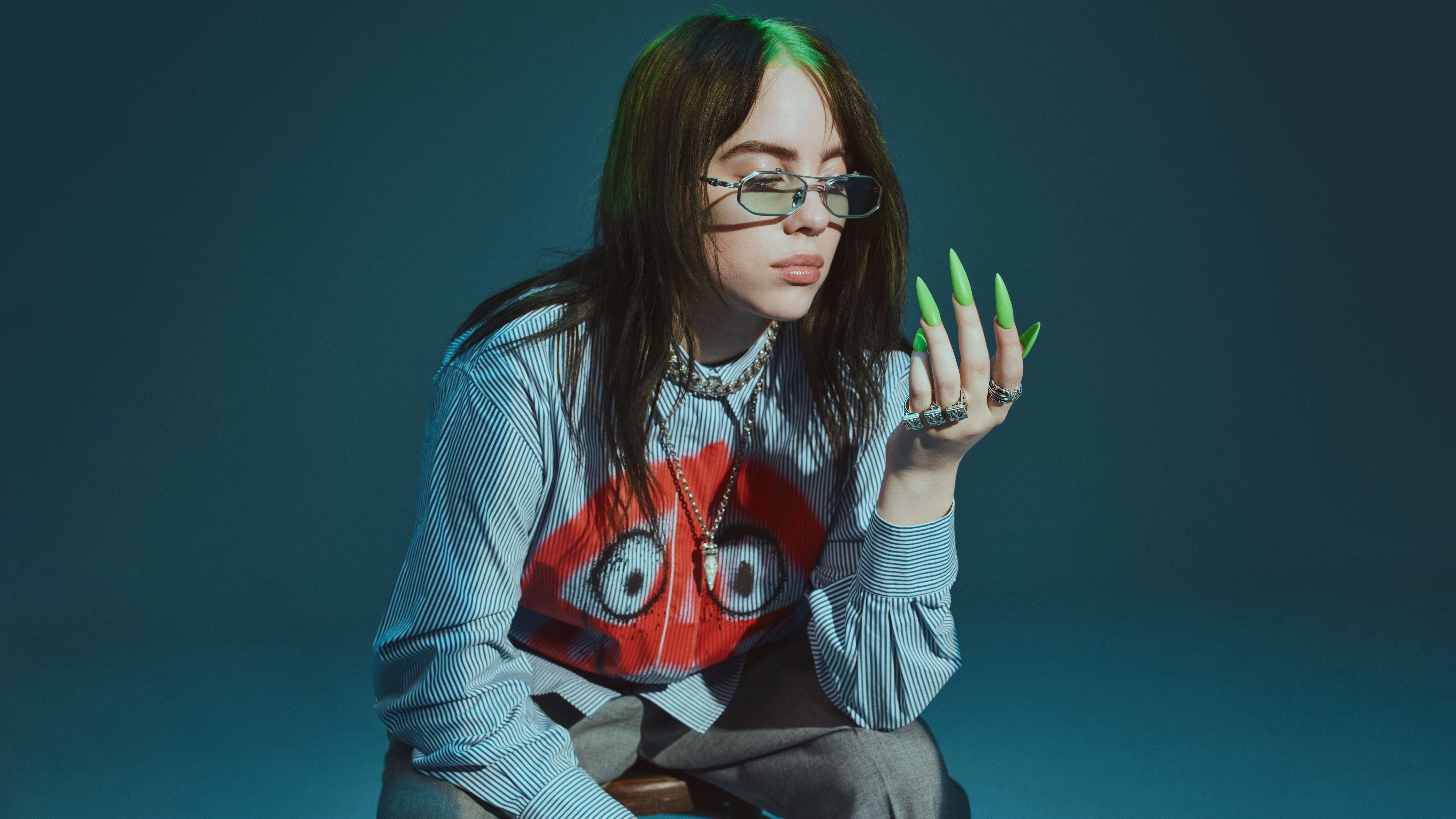 怪奇比莉 4K 桌布 壁紙 — Billie Eilish 戴著眼鏡，塗上螢光綠色指甲，身穿印有紅色卡通臉的條紋襯衫，在憂鬱的深藍綠色背景前擺姿勢。
