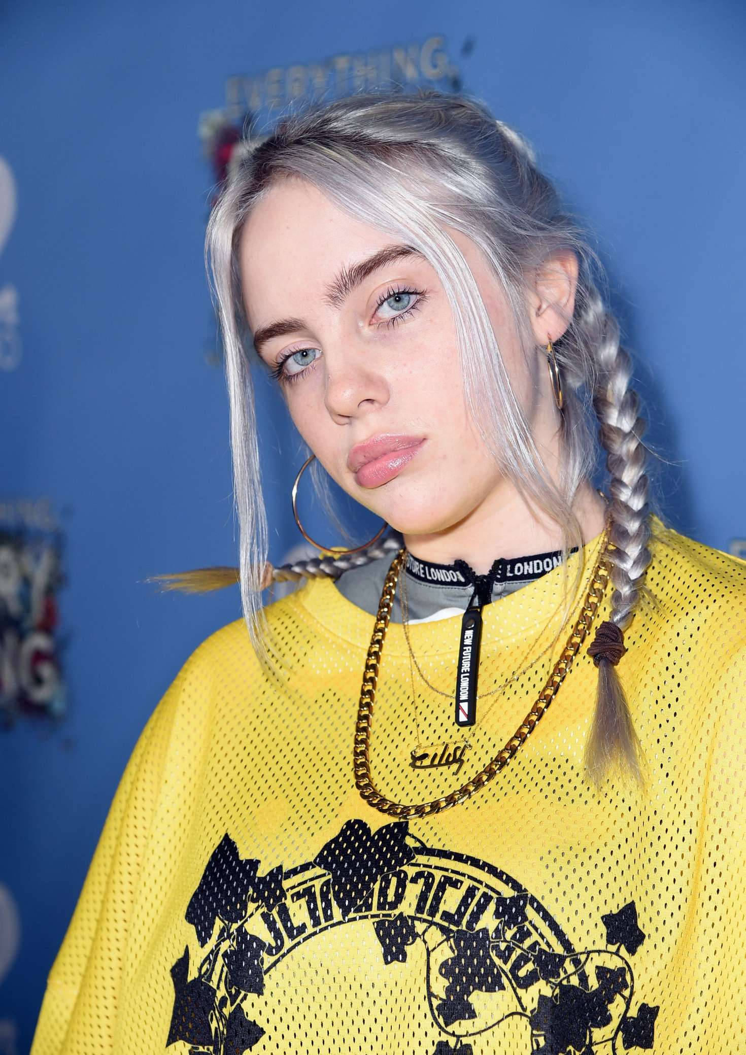 怪奇比莉 桌布 壁紙 — Billie Eilish 留著銀色辮子，戴著圈狀耳環，身穿亮黃色網眼上衣，背景為藍色。