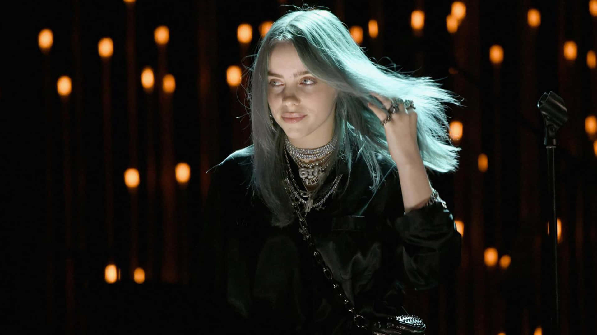 Billie Eilish Laptop 桌布 壁紙 — Billie Eilish 留著銀色頭髮，戴著多層項鍊，在溫暖的燈光下站在舞台上。