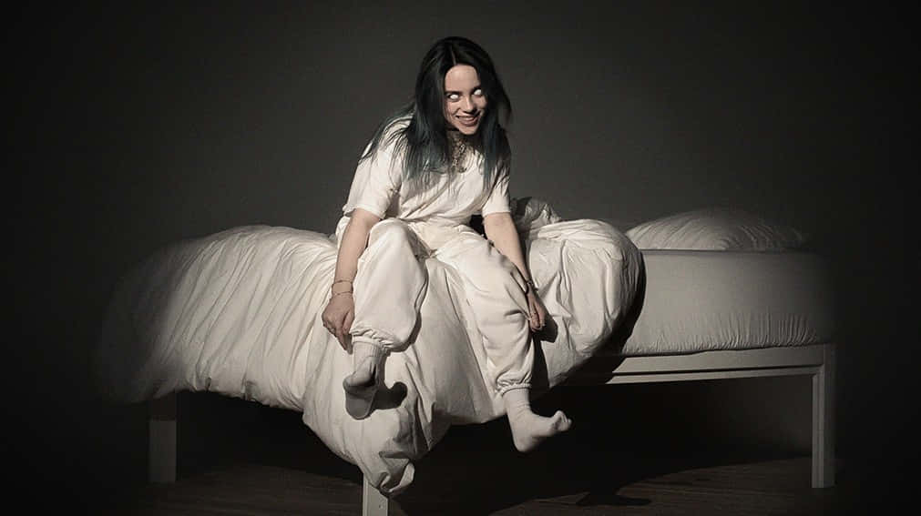 Billie Eilish Laptop 桌布 壁紙 — Billie Eilish 盤腿坐在光線昏暗的房間床上，身穿白色休閒服，對著鏡頭溫柔微笑。