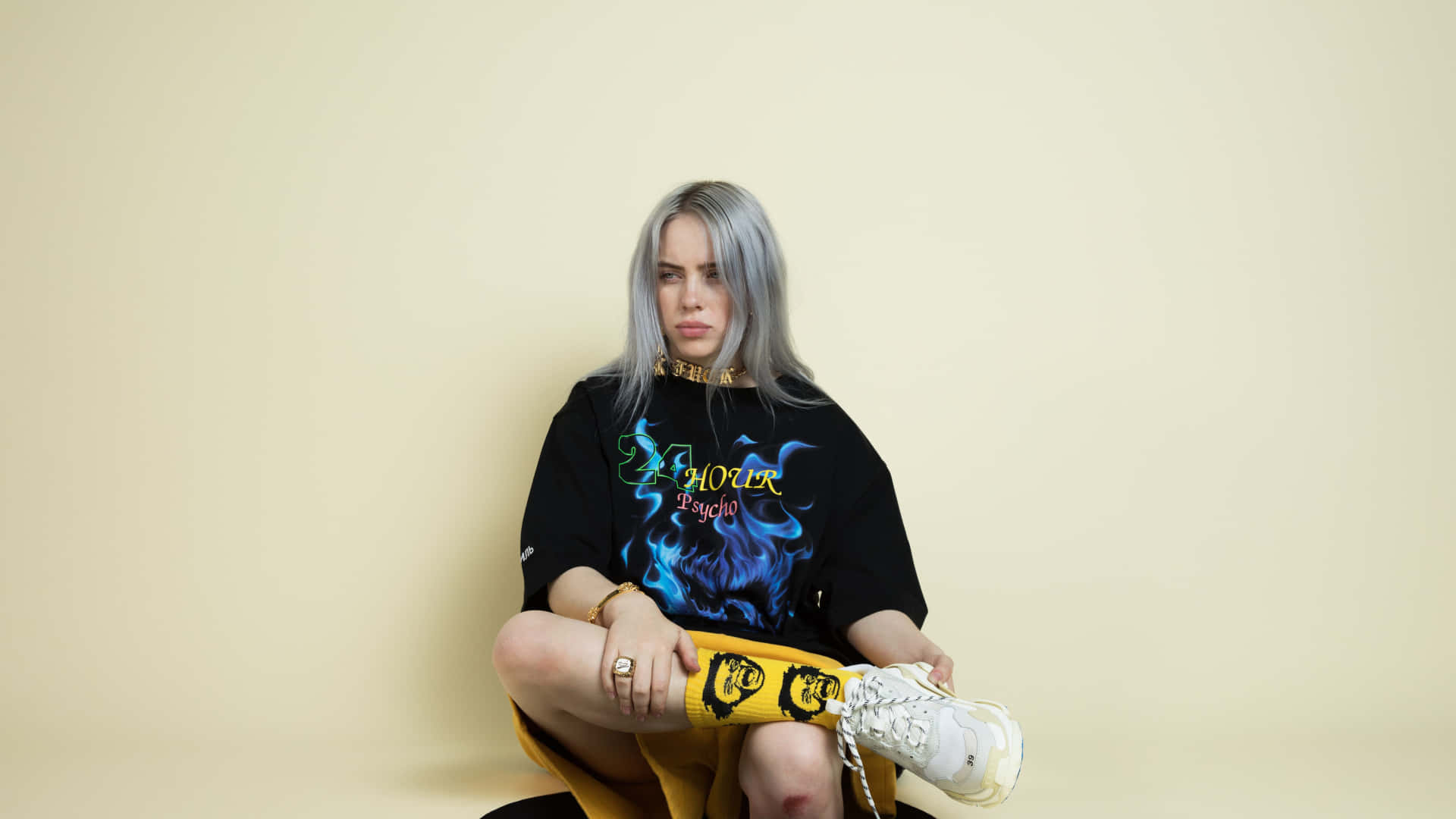 Billie Eilish Laptop 桌布 壁紙 — Billie Eilish 盤腿坐在純色米色牆前，身穿黑色圖案T恤、黃色褲子和白色運動鞋。