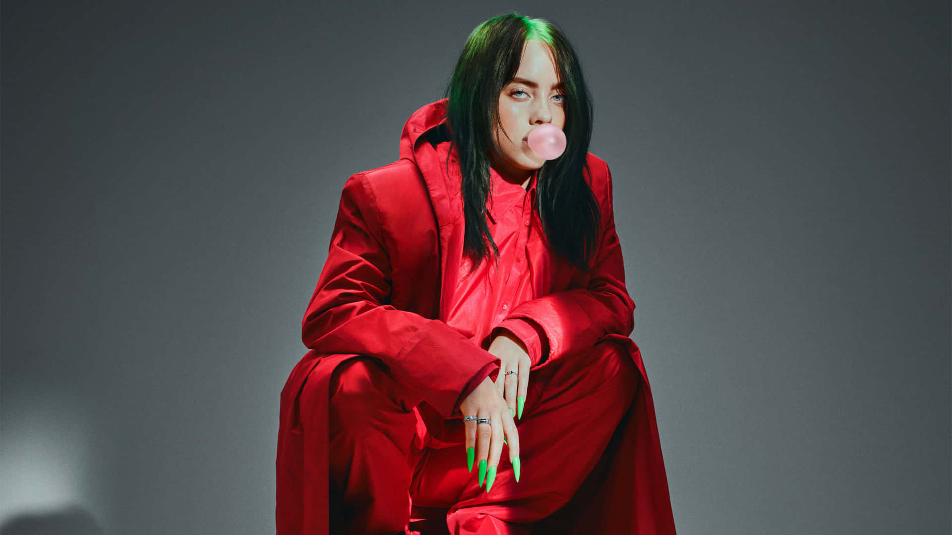 Billie Eilish Laptop 桌布 壁紙 — Billie Eilish 穿著大膽的紅色服裝坐著，對著柔和的灰色背景吹泡泡糖。