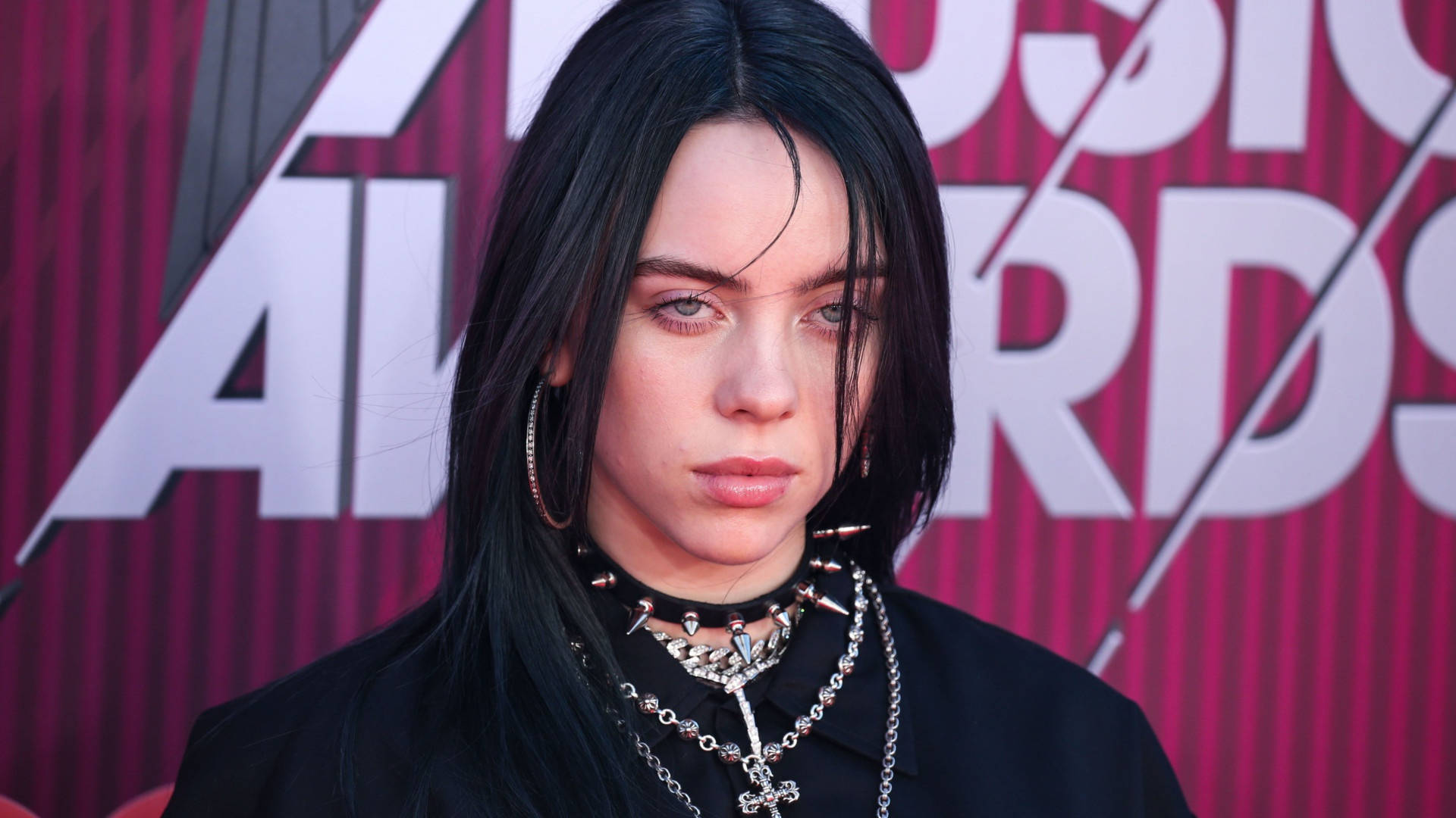 怪奇比莉 4K 桌布 壁紙 — Billie Eilish 站在紅地毯上，戴著多層鏈條和圈形耳環，背景是鮮明的粉紅色和白色「Music Awards」。