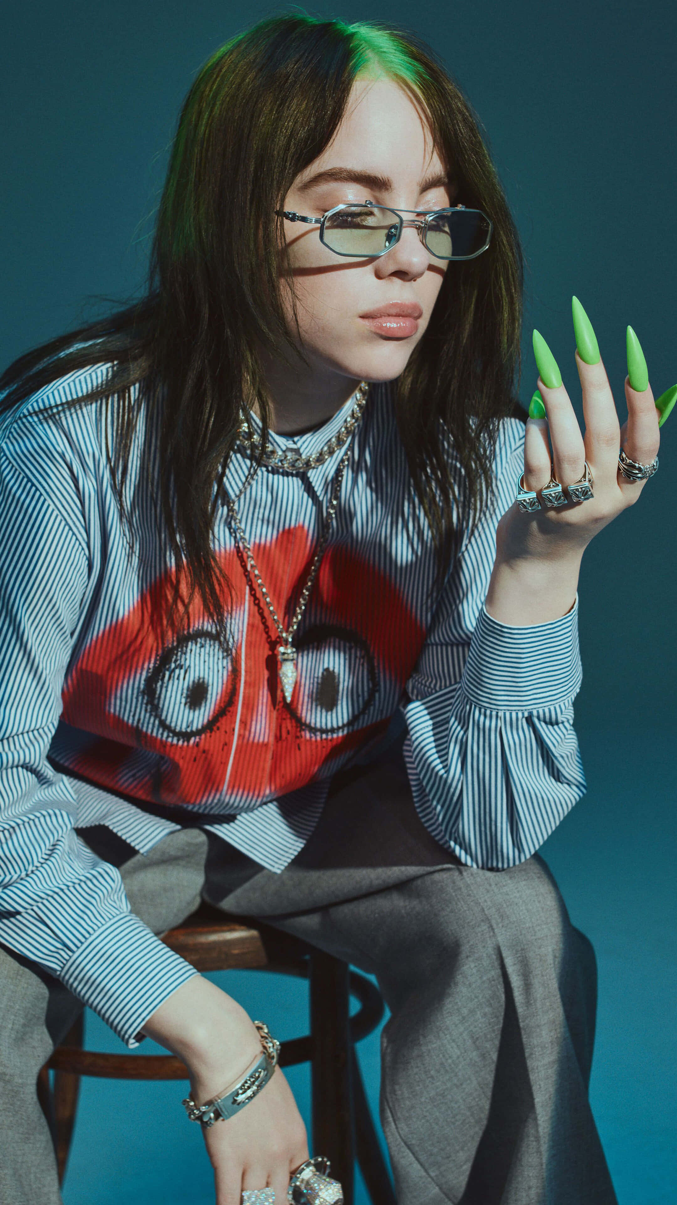 怪奇比莉 桌布 壁紙 — Billie Eilish 自信地坐在凳子上，戴著眼鏡和螢光綠色指甲，背景是藍綠色。