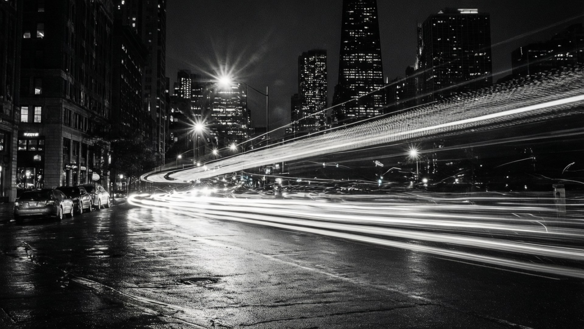 Night City Skyline 壁紙 — 黑白長曝光捕捉到濕漉漉的城市街道上夜間汽車燈光的軌跡，背景是高聳的摩天大樓。