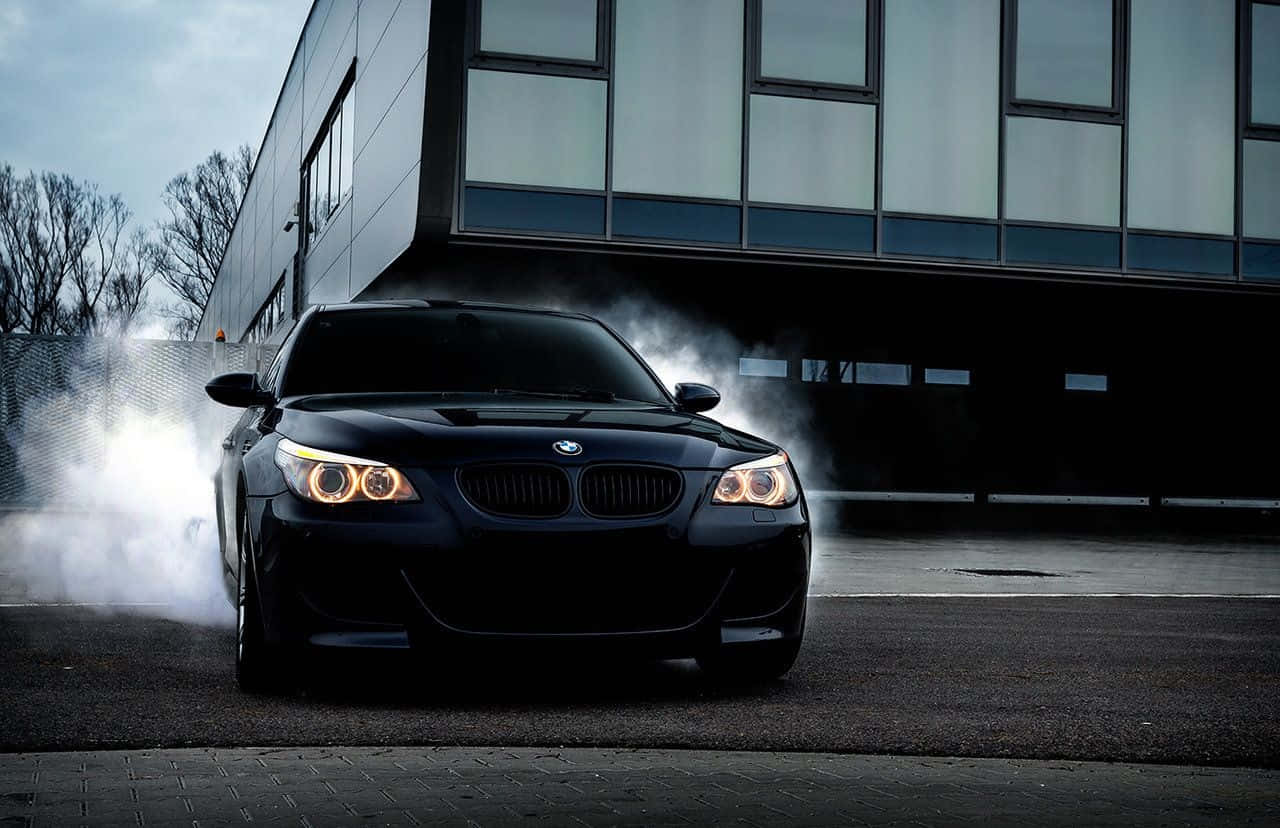 Bmw M5 桌布 壁紙 — 一輛黑色BMW跑車在陰天下一棟現代玻璃建築前甩尾，輪胎冒出滾滾濃煙。