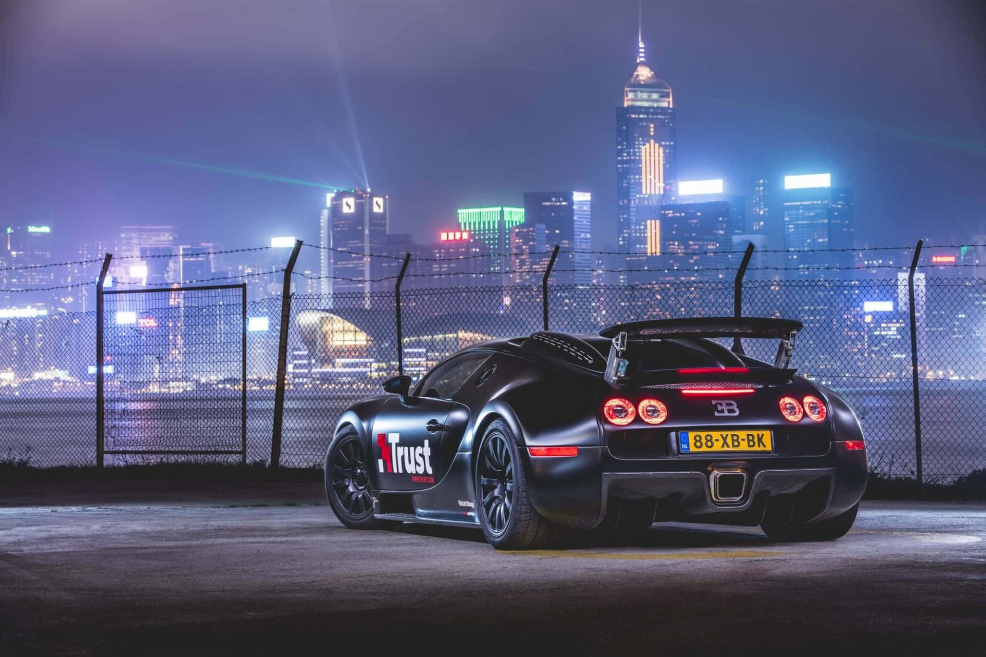 Bugatti Veyron 桌布 壁紙 — 一輛黑色Bugatti跑車，帶有發光的紅色尾燈，夜間停在鏈條圍欄後面燈火通明的城市天際線前。