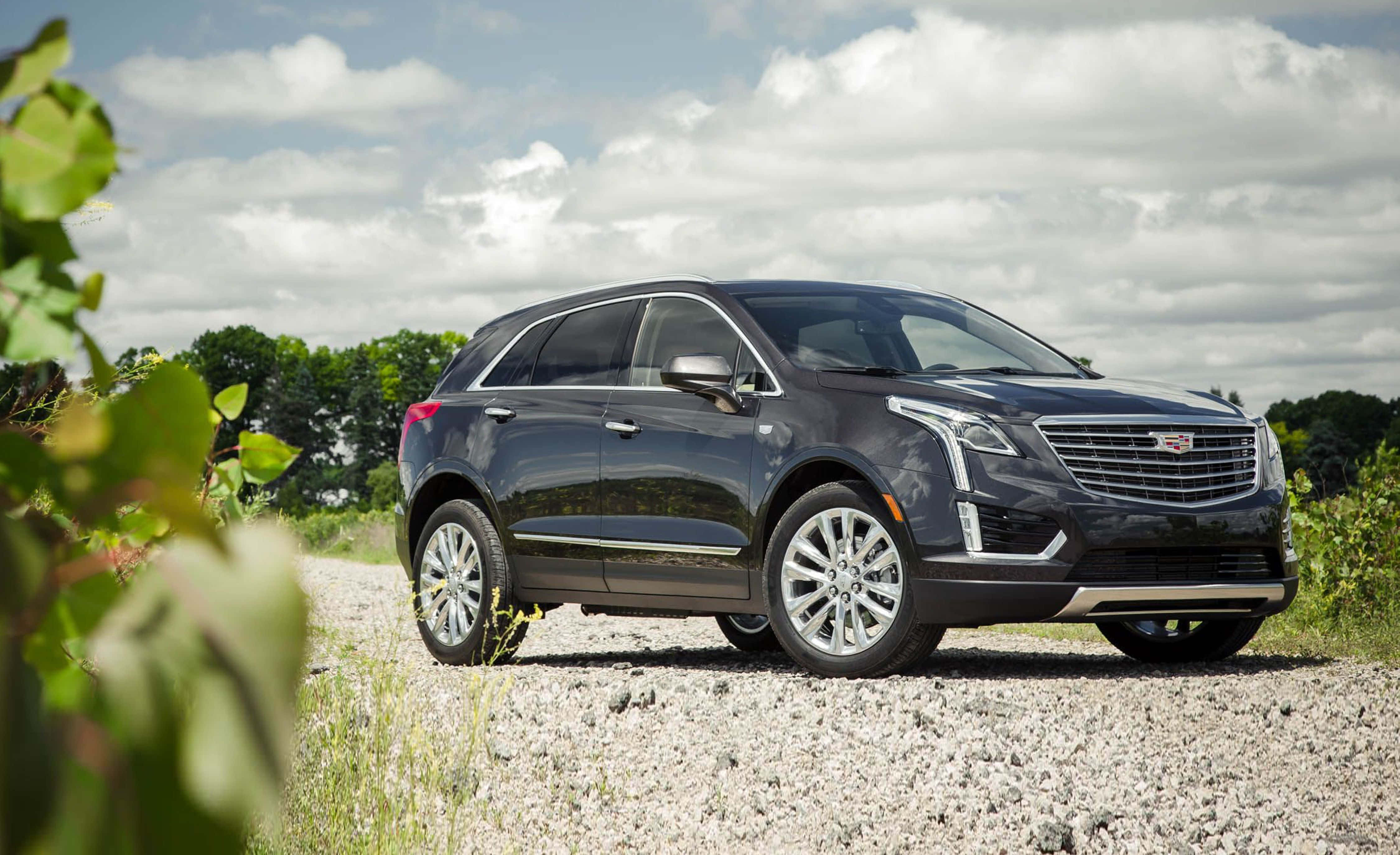 Cadillac Xt5 桌布 壁紙 — 一輛時尚的黑色凱迪拉克SUV停在碎石路上，背景有綠色樹葉和多雲的天空。