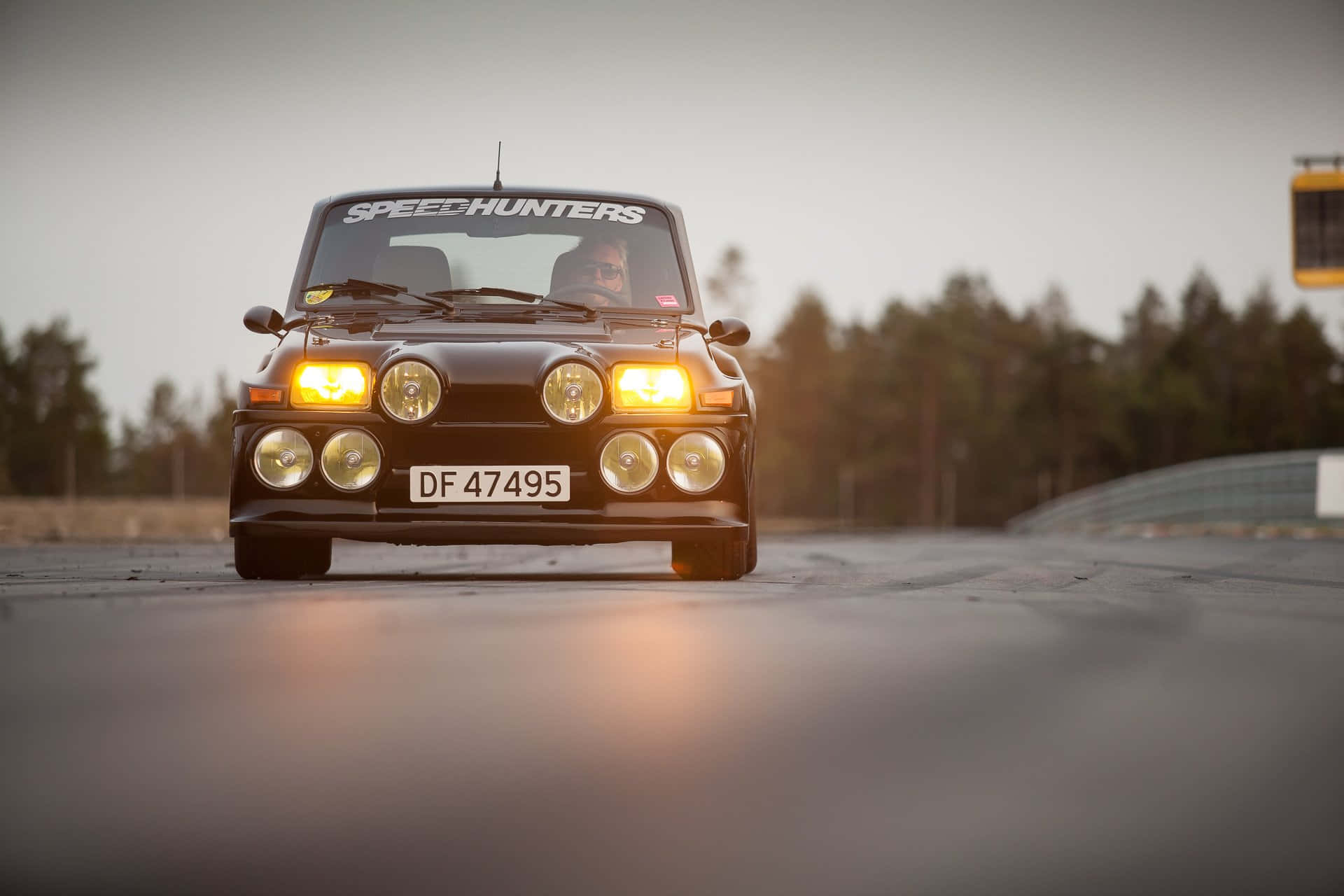 Renault 5 Turbo 桌布 壁紙 — 一輛黑色經典賽車，帶有發光的車頭燈，在黃昏時面向賽道前方，背景有樹木。