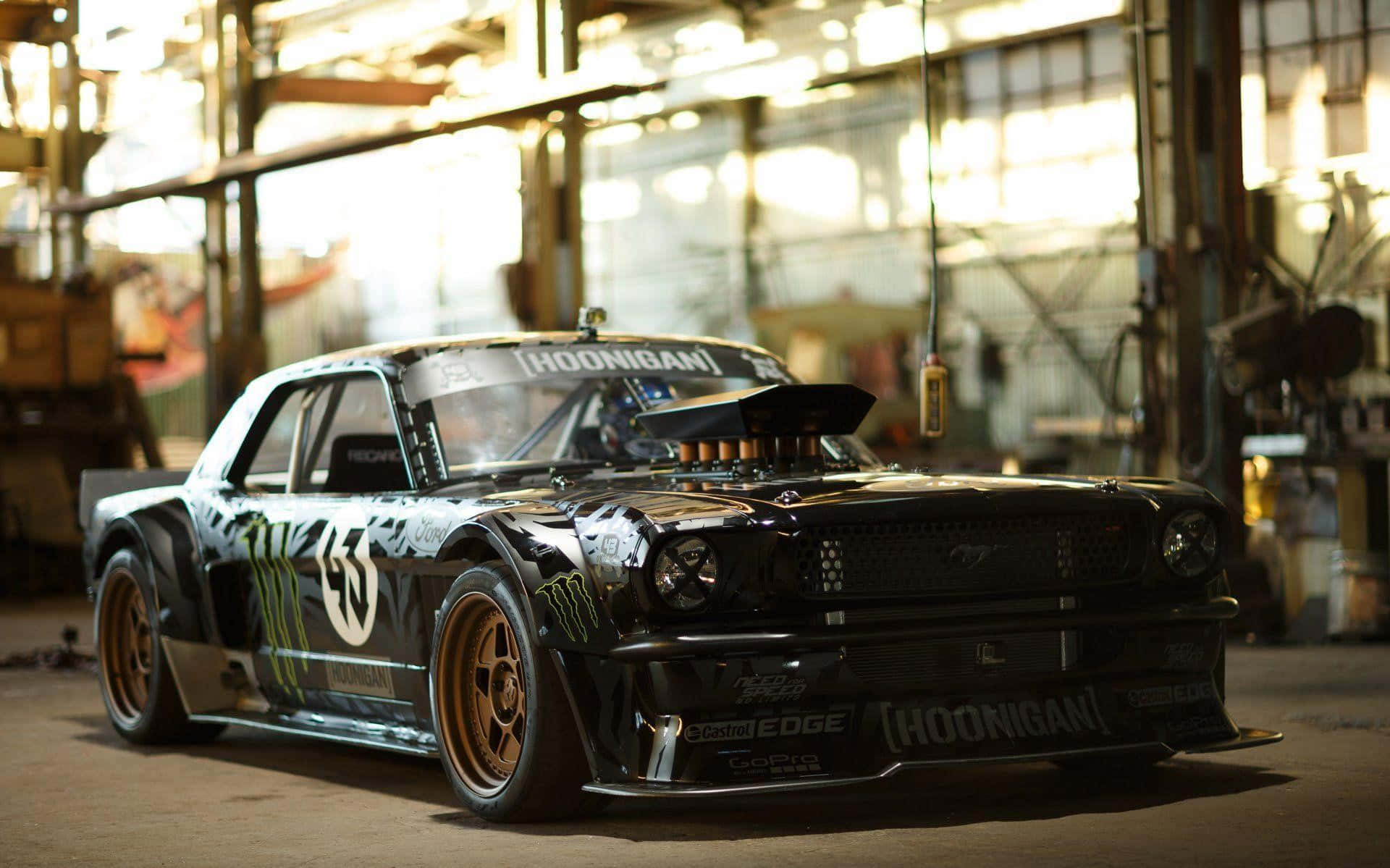 Hoonigan 桌布 壁紙 — 一輛黑色Hoonigan Mustang甩尾車配金色輪圈，停放在陽光照射的工業車庫內。