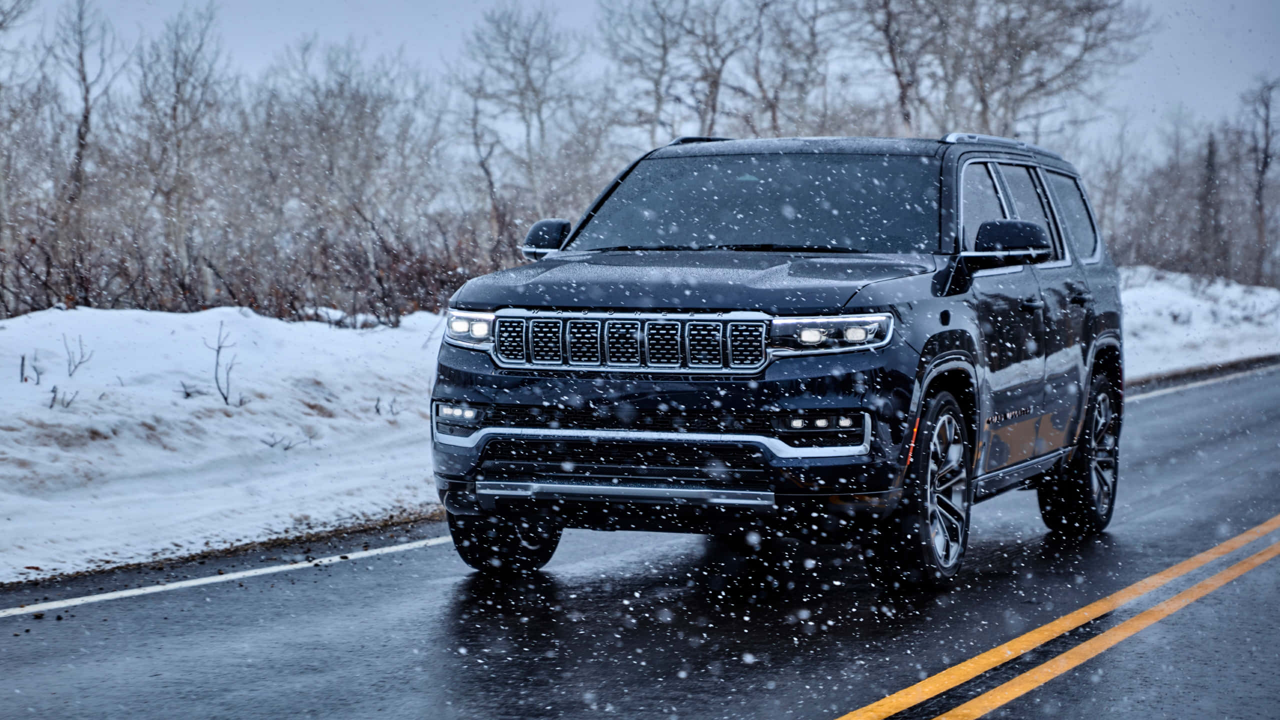 Jeep Wagoneer 桌布 壁紙 — 一輛黑色Jeep Grand Wagoneer在濕滑的道路上行駛，穿過飄落的雪花，兩旁是積雪的樹木。