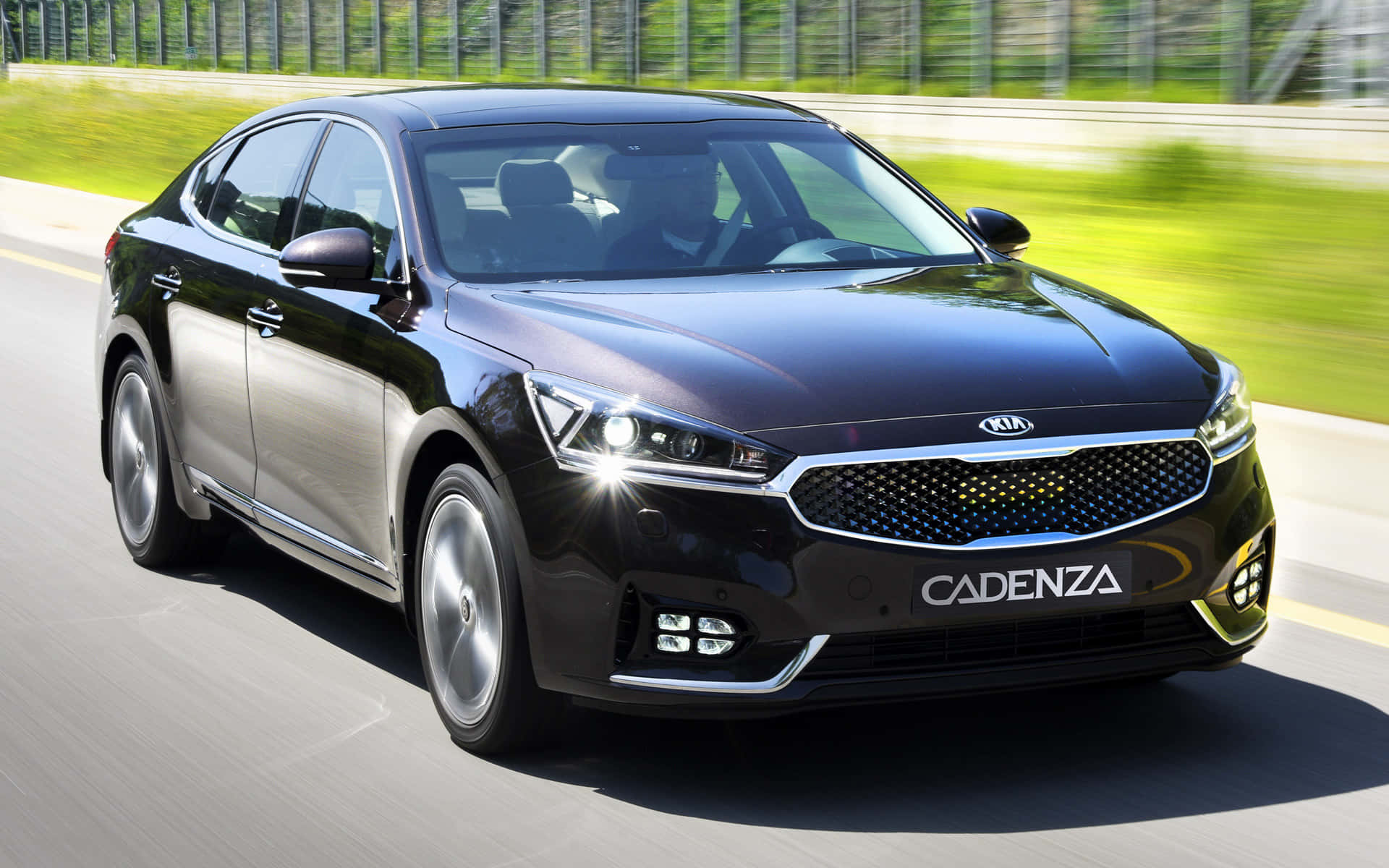 Kia Cadenza 桌布 壁紙 — 一輛時尚的黑色Kia Cadenza轎車在陽光明媚的高速公路上疾馳，背景是模糊的綠色植物。