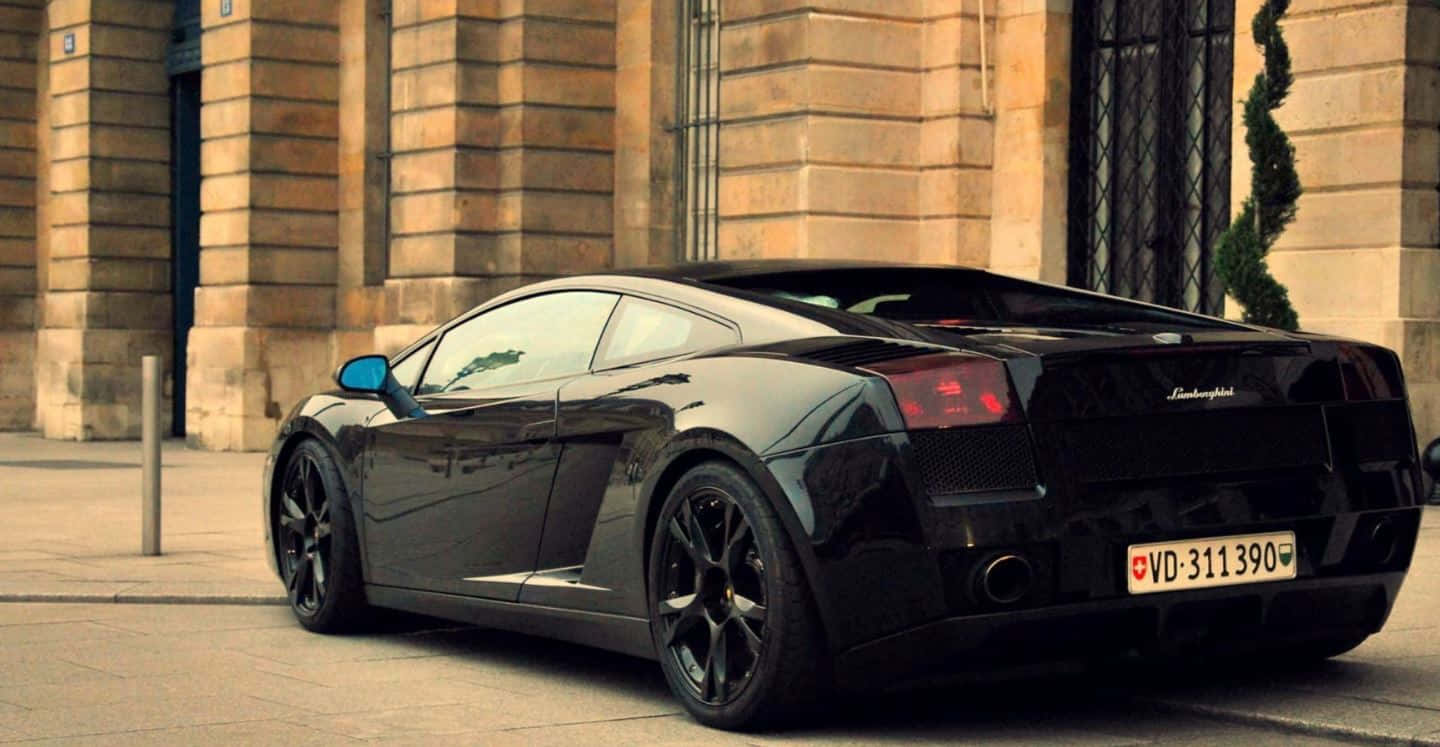 Lamborghini Gallardo 桌布 壁紙 — 一架時尚黑色藍寶堅尼Gallardo停放在陽光照射的石頭人行道上，旁邊是一座宏偉的古典建築。