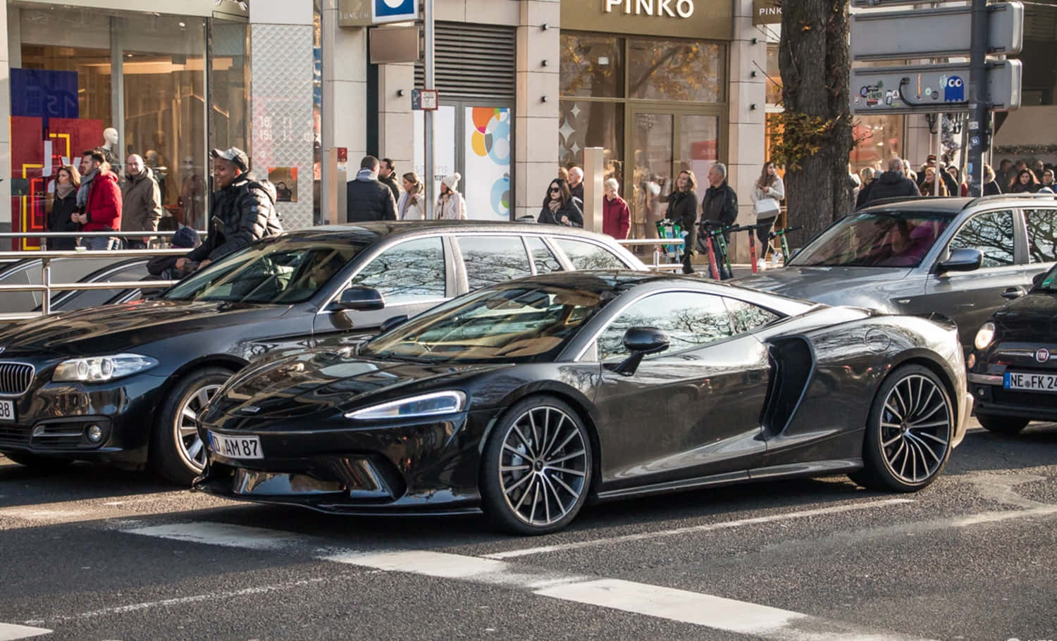 Mclaren Gt 桌布 壁紙 — 一輛時尚的黑色McLaren跑車在熙來攘往的街道上滑行，街道兩旁盡是行人和商店。