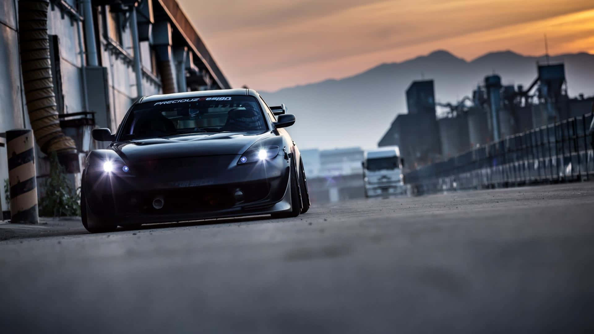 Mazda Rx-8 桌布 壁紙 — 一輛黑色改裝跑車帶有發光車頭燈，在日落時分沿著工業道路駛向鏡頭，背景有山脈。