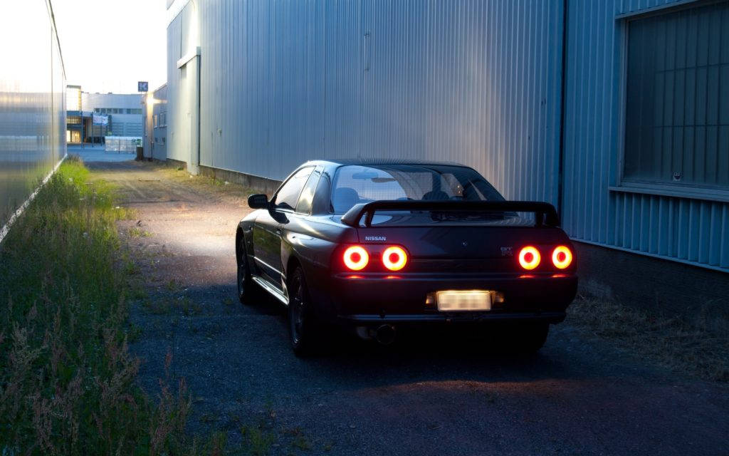 黑色跑車帶有發光尾燈，在黃昏時停泊在藍色倉庫旁。 — 城市天際線汽車 壁紙