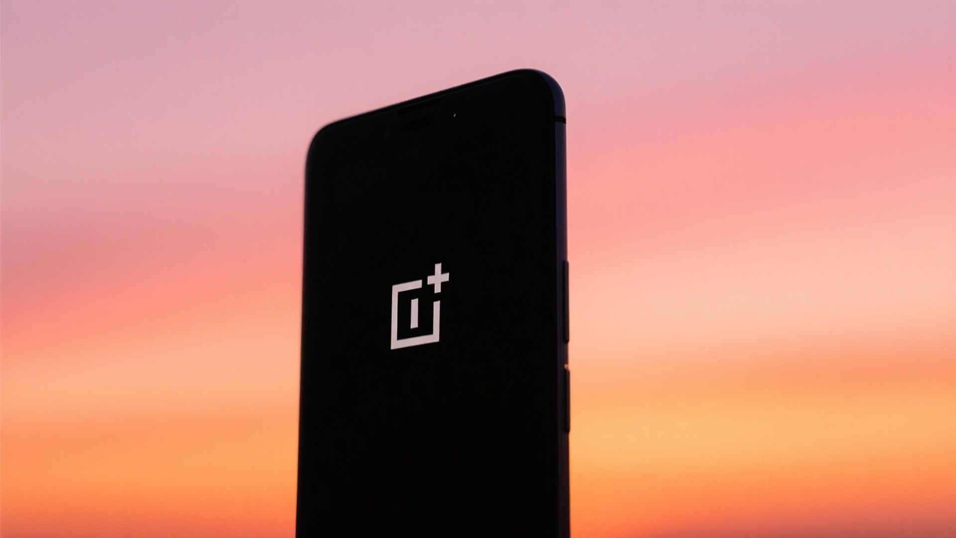 Sunset Phone Silhouette 壁紙 — 一部黑色 OnePlus 智慧型手機直立於充滿活力的日落天空前，混合了粉紅、橙和蜜桃色調。