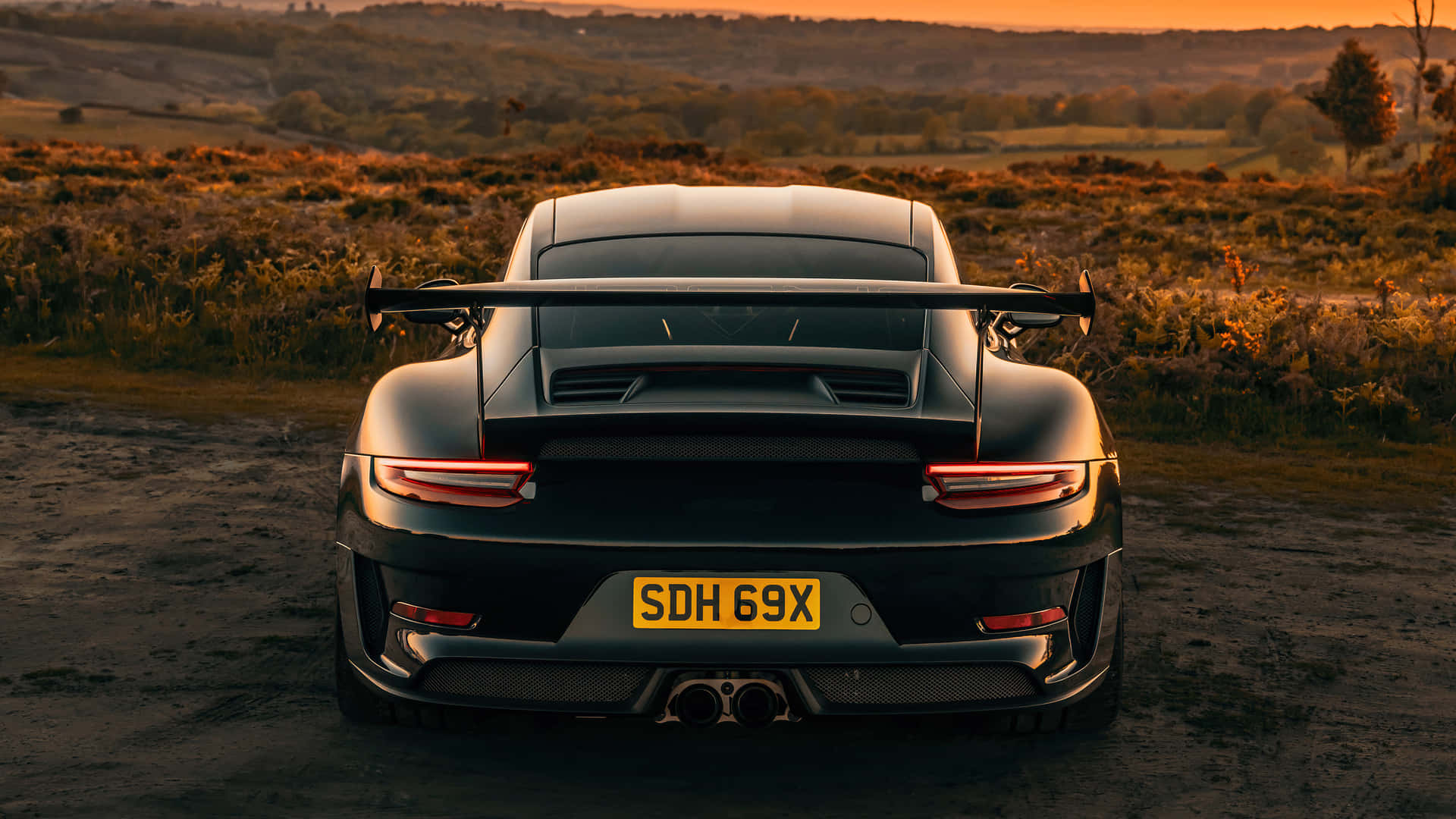  壁紙 — 一輛黑色保時捷911 GT2 RS停放在日落時分的泥土路上，金色光線照亮了背後崎嶇的鄉村景色。