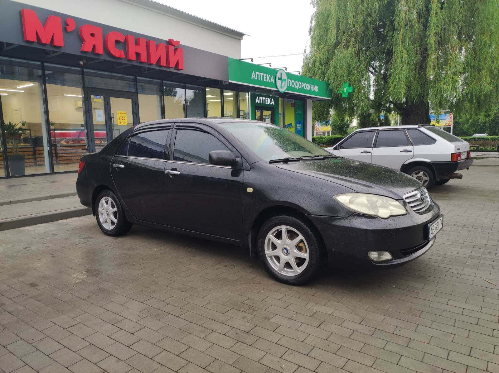 Byd F3 桌布 壁紙 — 一輛黑色轎車停泊在潮濕的磚砌路面上，前面是肉店和藥房，頭頂有綠樹。