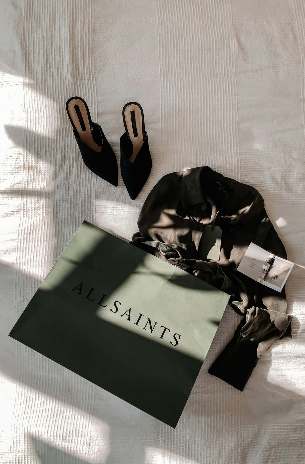  壁紙 — 黑色天鵝絨穆勒鞋和深色襯衫放在AllSaints包包旁邊，位於陽光照射的白色床罩上，帶有條紋質感。
