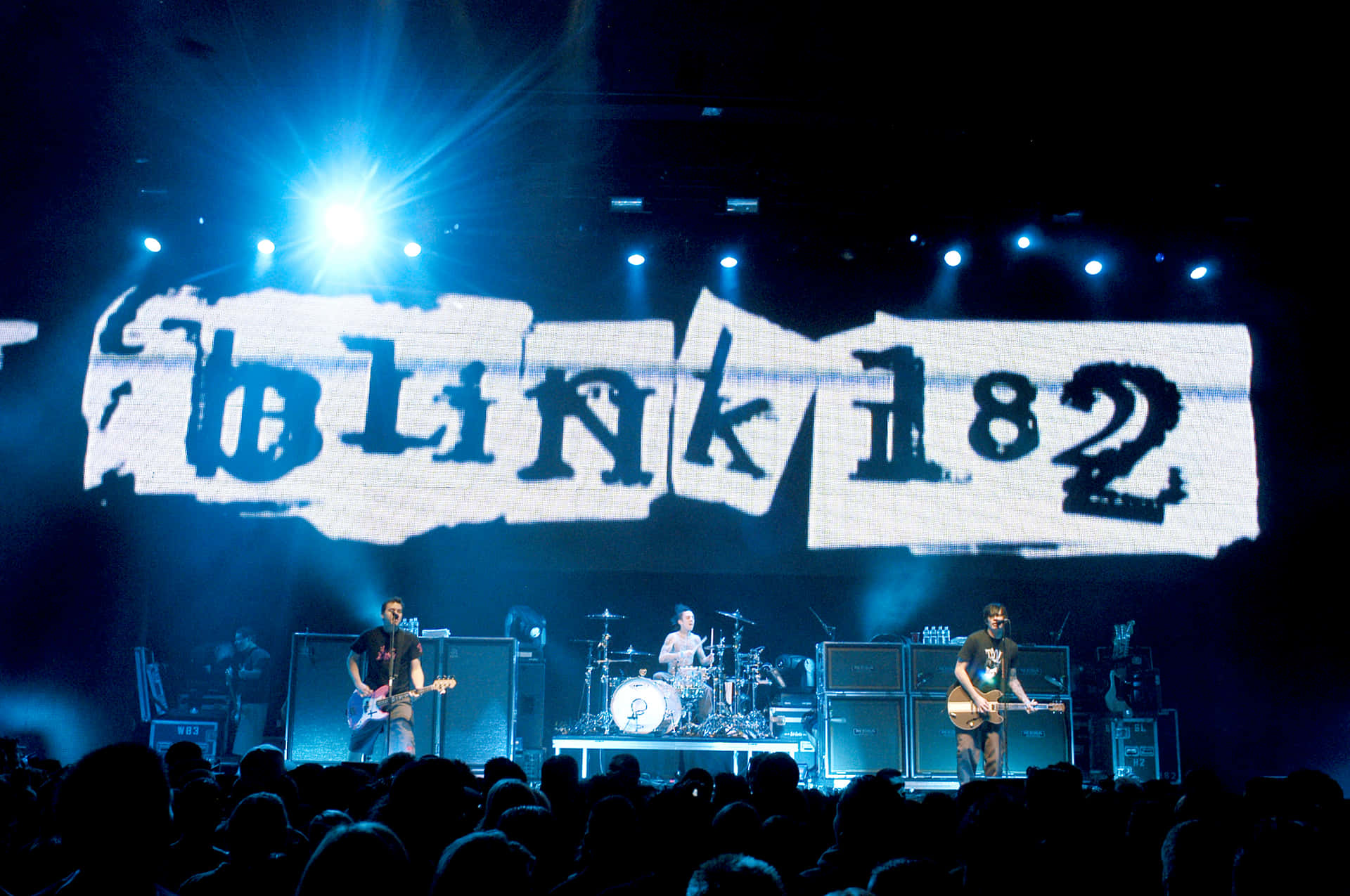  壁紙 — Blink 182 樂團在黑暗的舞台上進行現場表演，舞台沐浴在藍色燈光中，身後有一個巨大的撕紙樂團標誌。