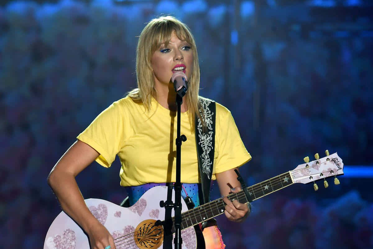 Taylor Swift Lover 桌布 壁紙 — 一位金髮女子身穿黃色襯衫，在藍色舞台燈光下，一邊彈奏心形圖案的原聲吉他，一邊對著麥克風唱歌。