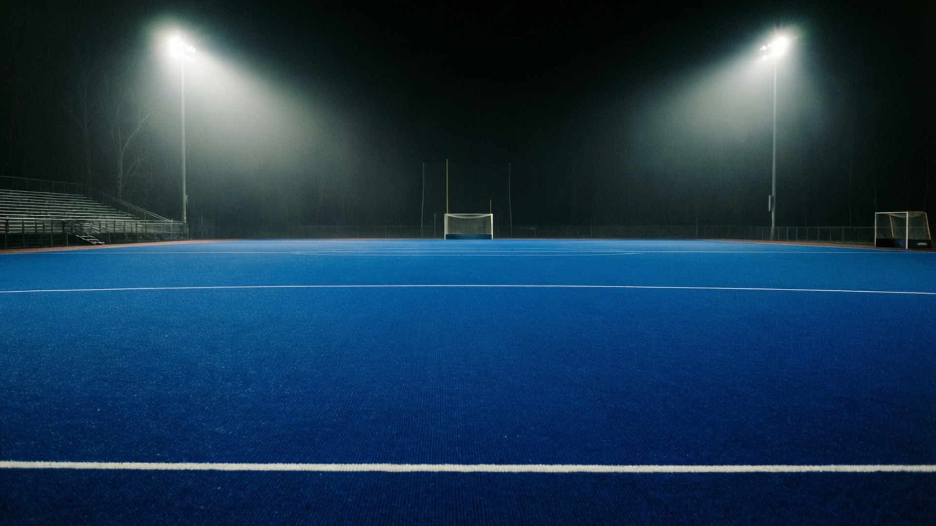 Empty Pitch Night Lights 壁紙 — 夜晚一個廣闊的藍色人造草地球場，由兩盞明亮的體育場燈光照亮，光束投射在空蕩的球門和看台上。
