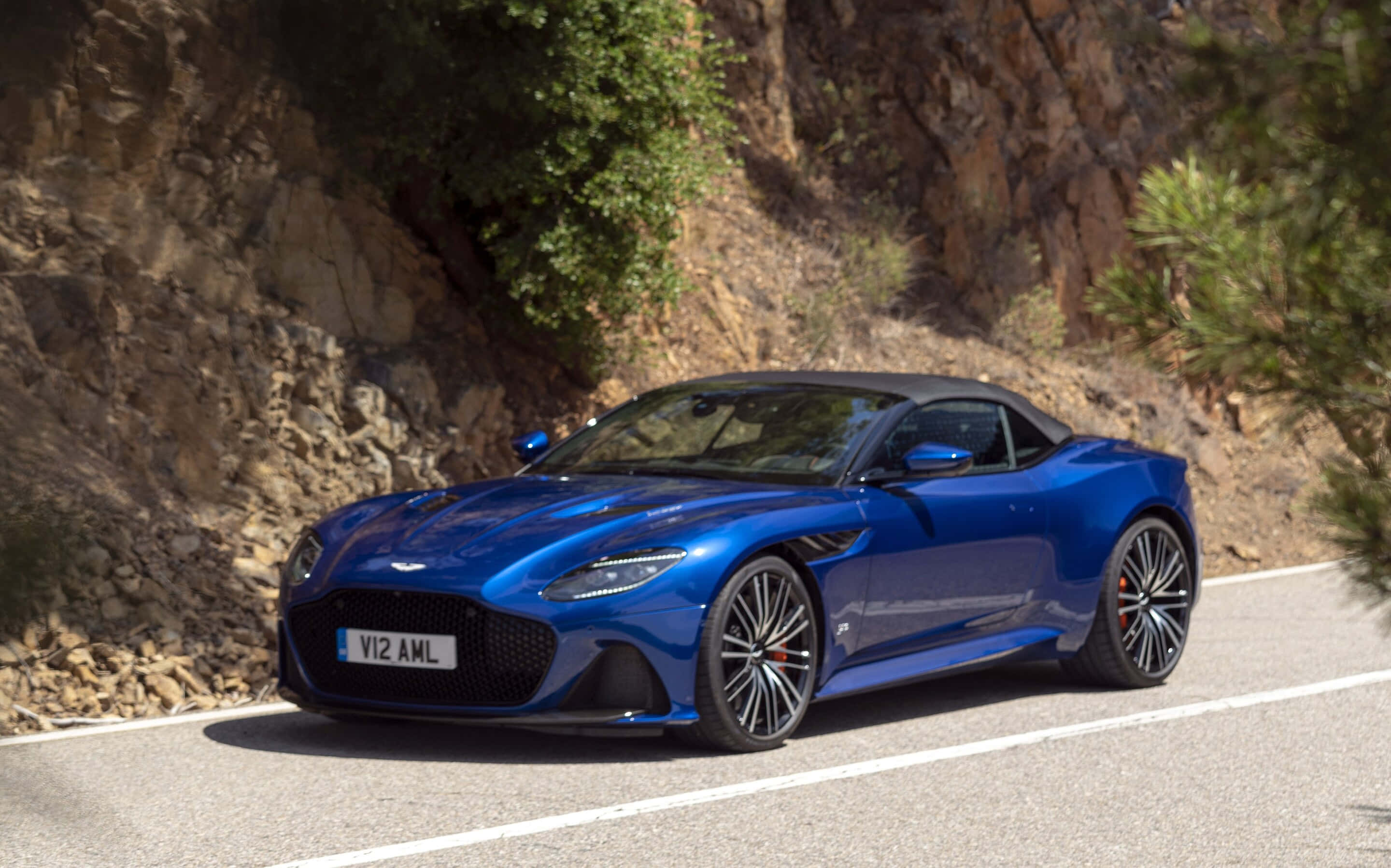 Aston Martin Dbs Superleggera 桌布 壁紙 — 一輛充滿活力的藍色 Aston Martin V12 Vantage 敞篷跑車沿著蜿蜒的山路巡航，兩旁是岩石峭壁和綠意。