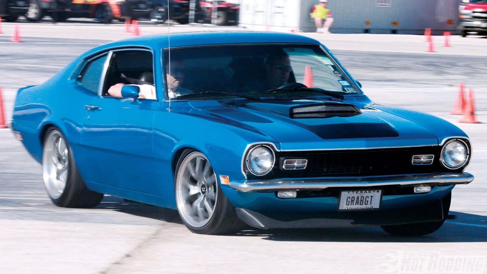 Ford Maverick 桌布 壁紙 — 一輛充滿活力的藍色經典肌肉車在鋪設的賽道上飛馳，周圍環繞著橙色交通錐。