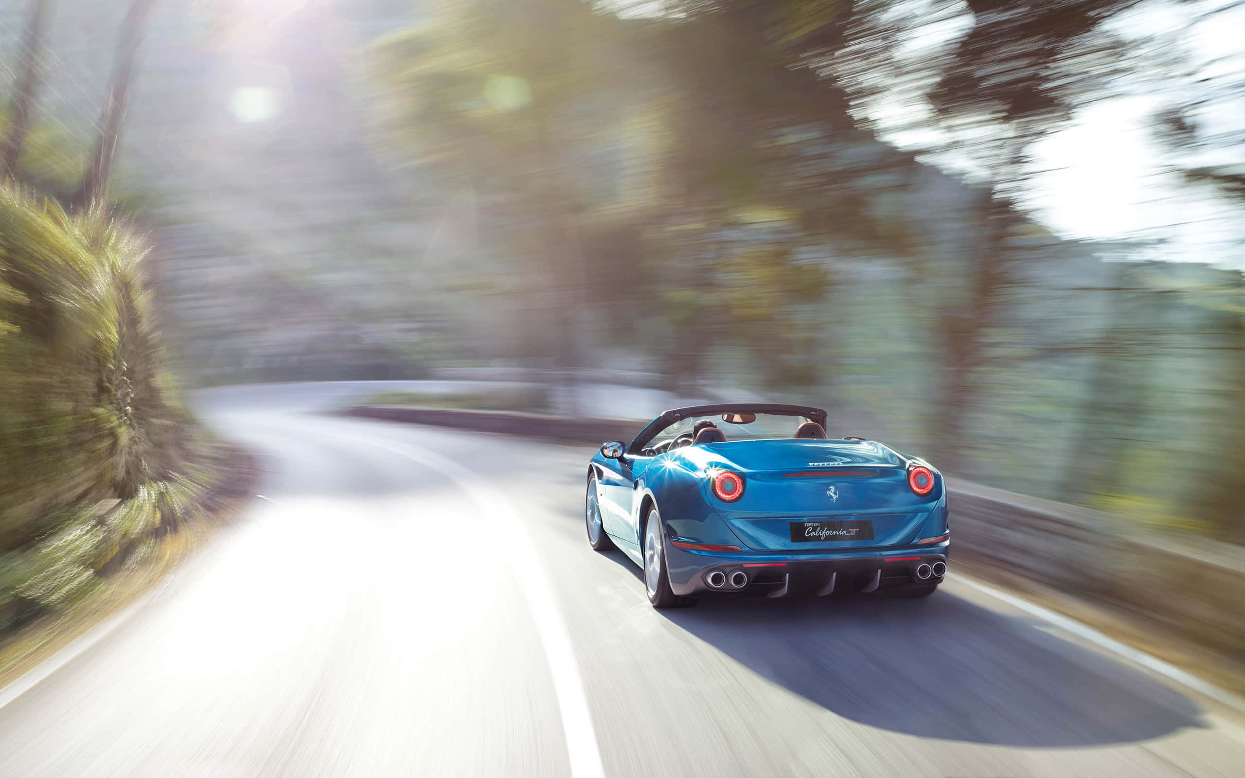 Ferrari California T 桌布 壁紙 — 一輛充滿活力的藍色法拉利 California T 在蜿蜒的林間道路上疾馳，帶有動態模糊和太陽眩光。