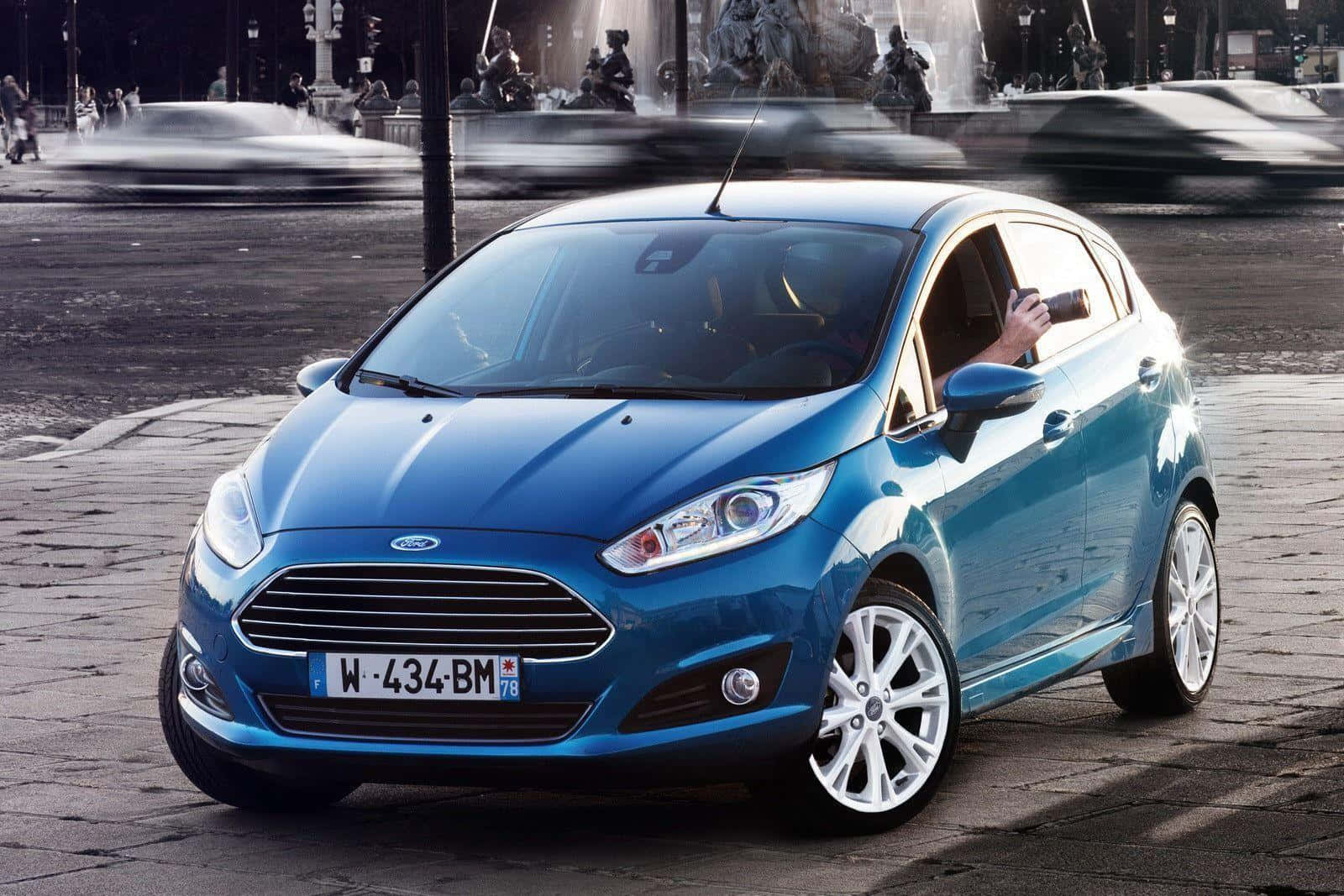 Ford Fiesta 桌布 壁紙 — 一輛鮮藍色的福特Fiesta停泊在鵝卵石街道上，背景有模糊的城市動態和一個噴泉。
