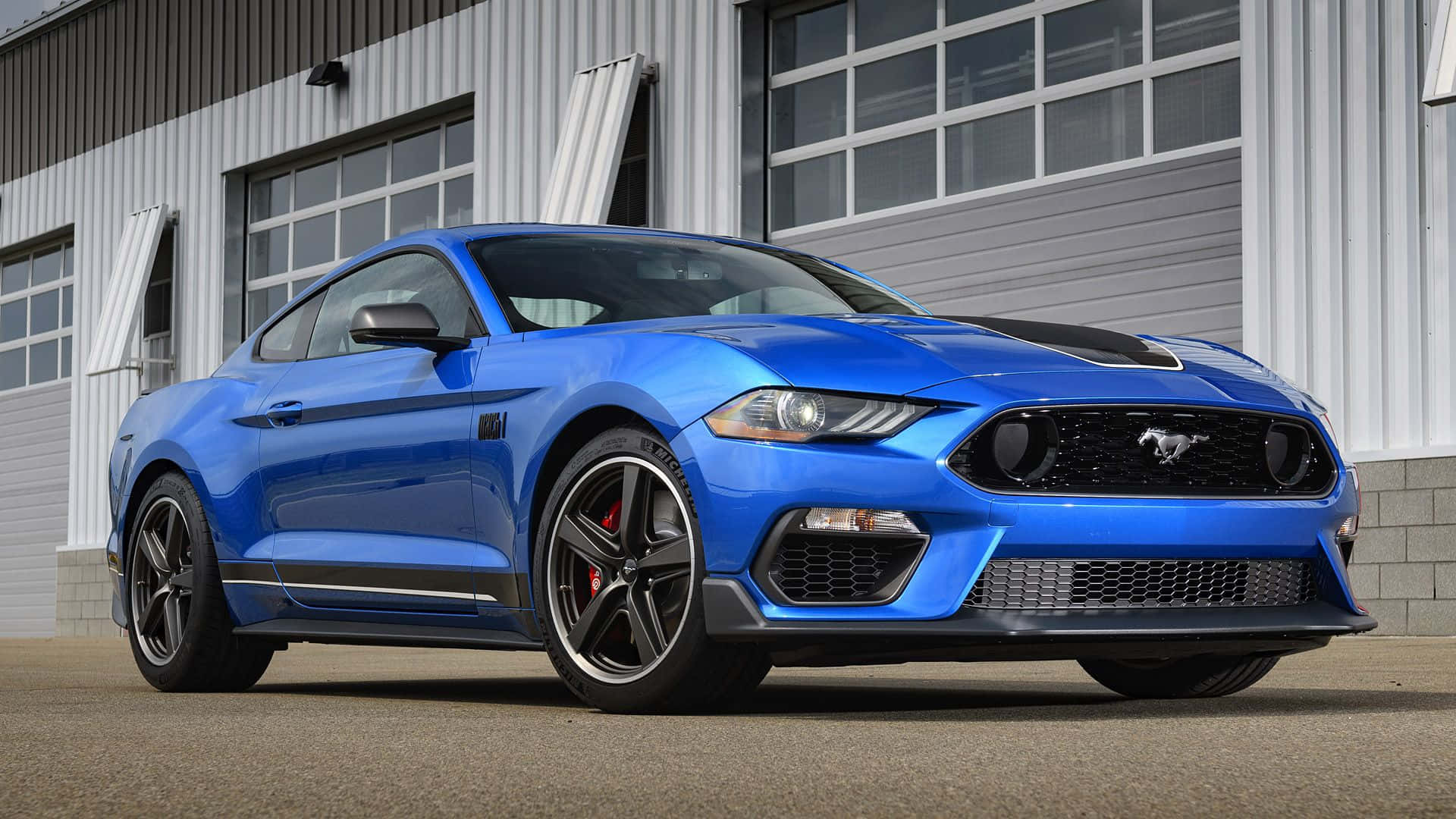 Ford Mustang Mach 1 桌布 壁紙 — 一輛充滿活力的藍色福特野馬跑車，帶有黑色飾邊，停在一棟現代工業建築前。