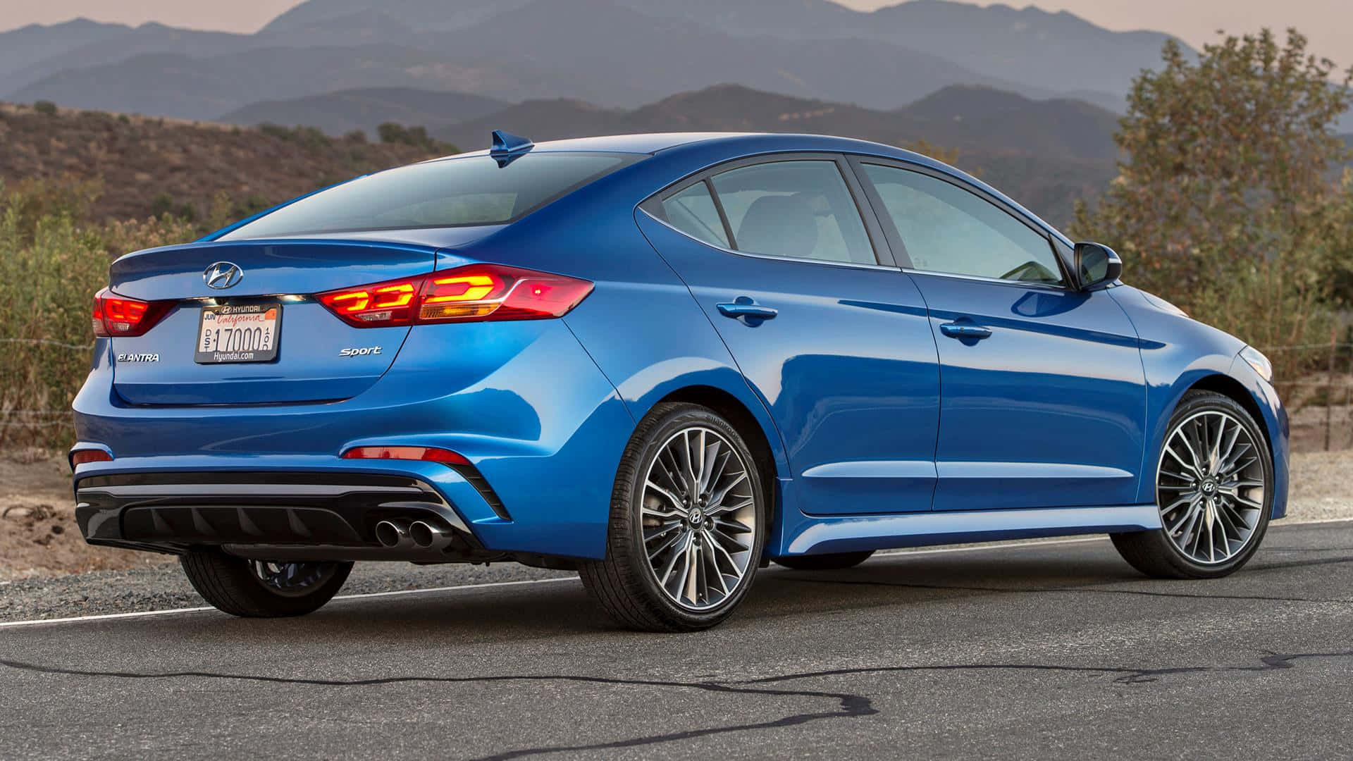 Hyundai Elantra 桌布 壁紙 — 一輛充滿活力的藍色現代 Elantra Sport 在黃金時段的光芒下閃耀，停泊在沙漠公路上，背景是山脈。