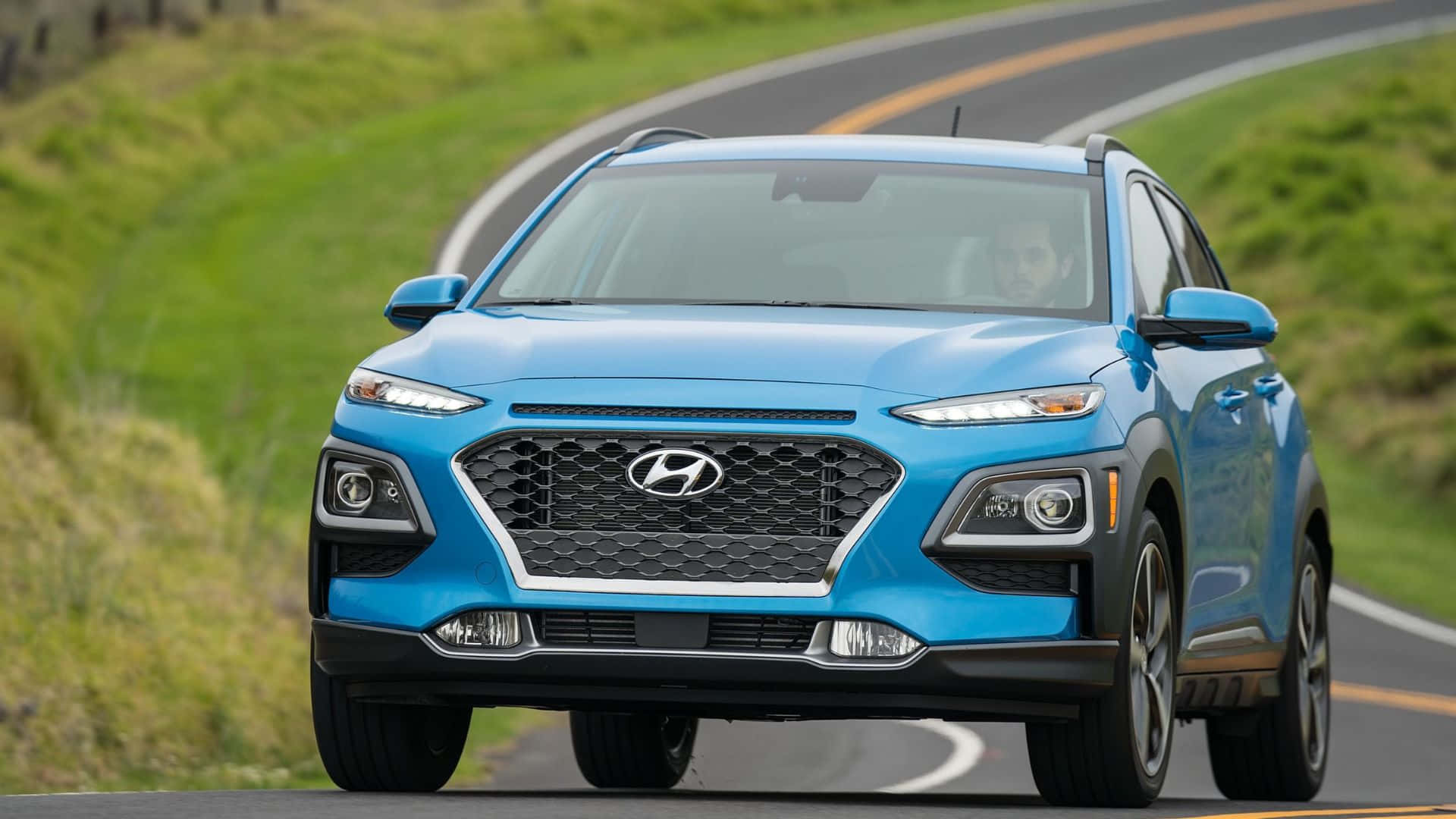 Hyundai Kona 桌布 壁紙 — 一輛鮮藍色的現代Kona SUV在明亮的日光下行駛在蜿蜒的瀝青路上，穿過鬱鬱蔥蔥的綠色山丘。