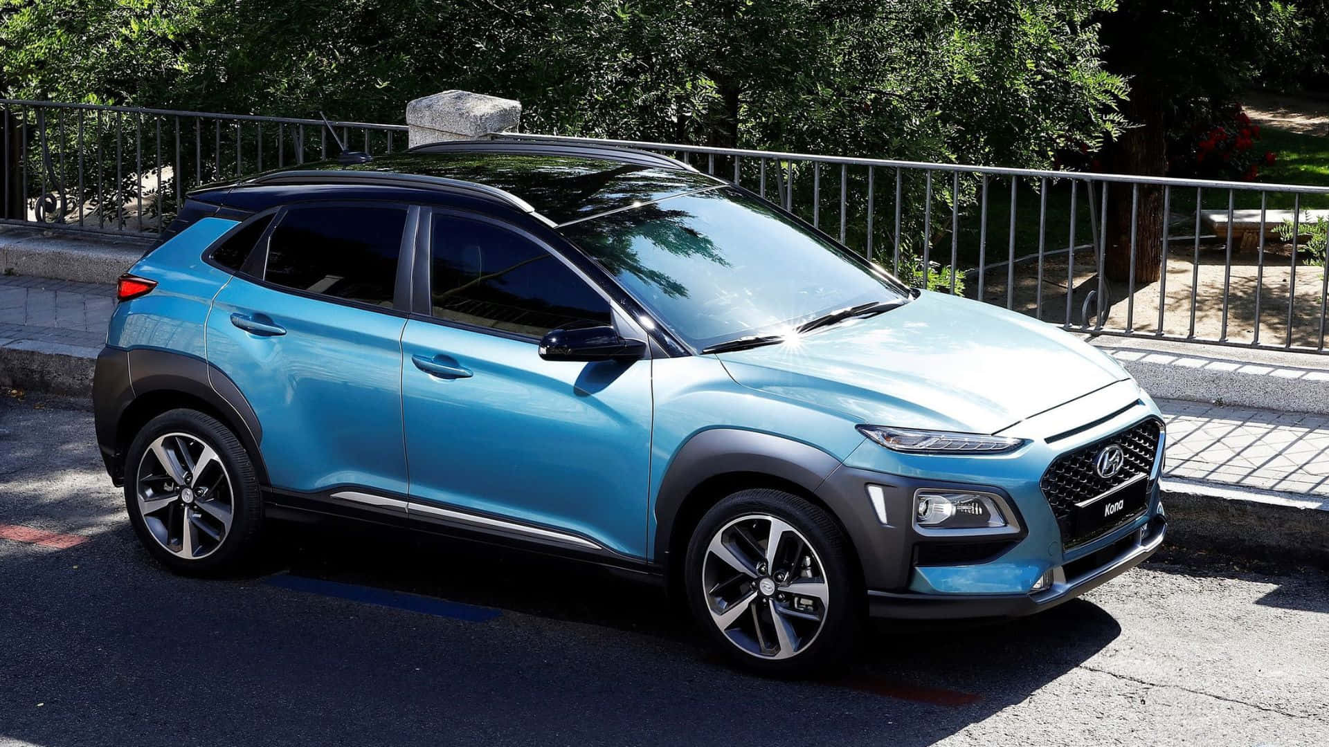 Hyundai Kona 桌布 壁紙 — 一輛鮮藍色的現代Kona SUV停放在陽光明媚的街道上，旁邊有金屬欄杆和綠樹。