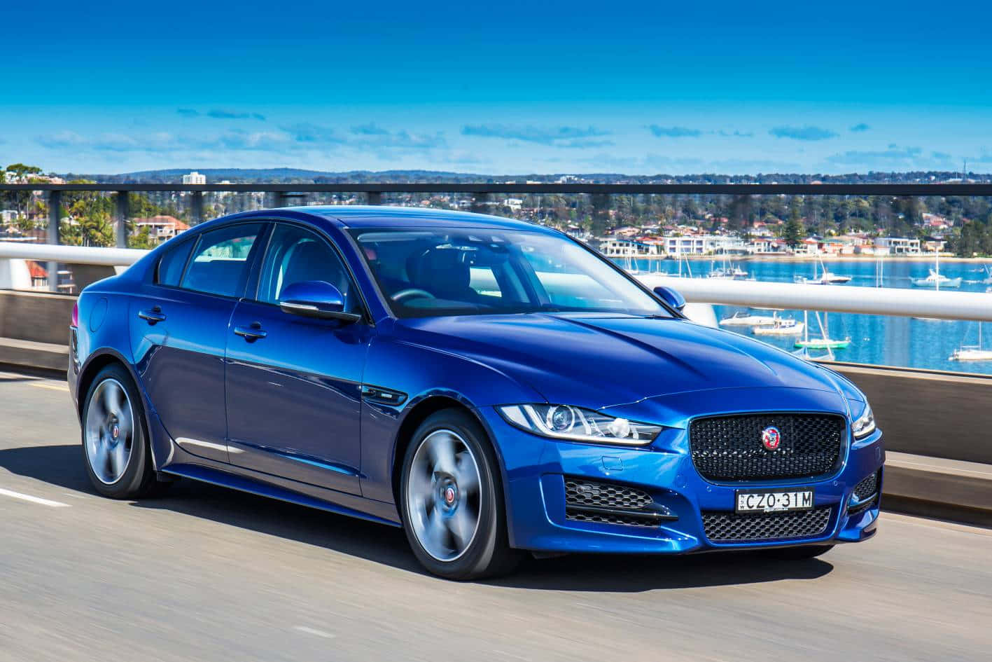 Jaguar Xe 桌布 壁紙 — 一輛充滿活力的藍色積架XF轎車在沿海公路上巡航，背景是碼頭和晴朗的藍天。