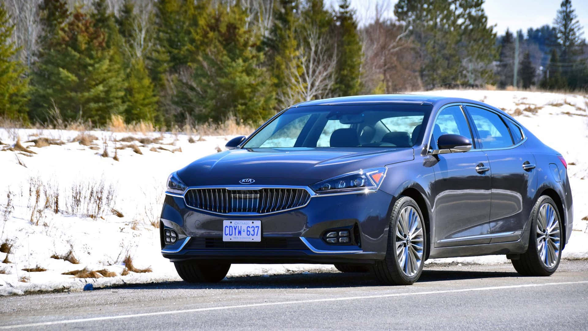 Kia Cadenza 桌布 壁紙 — 一輛時尚的藍色Kia轎車在明亮的日光下行駛在積雪的路邊，背景是常青樹。