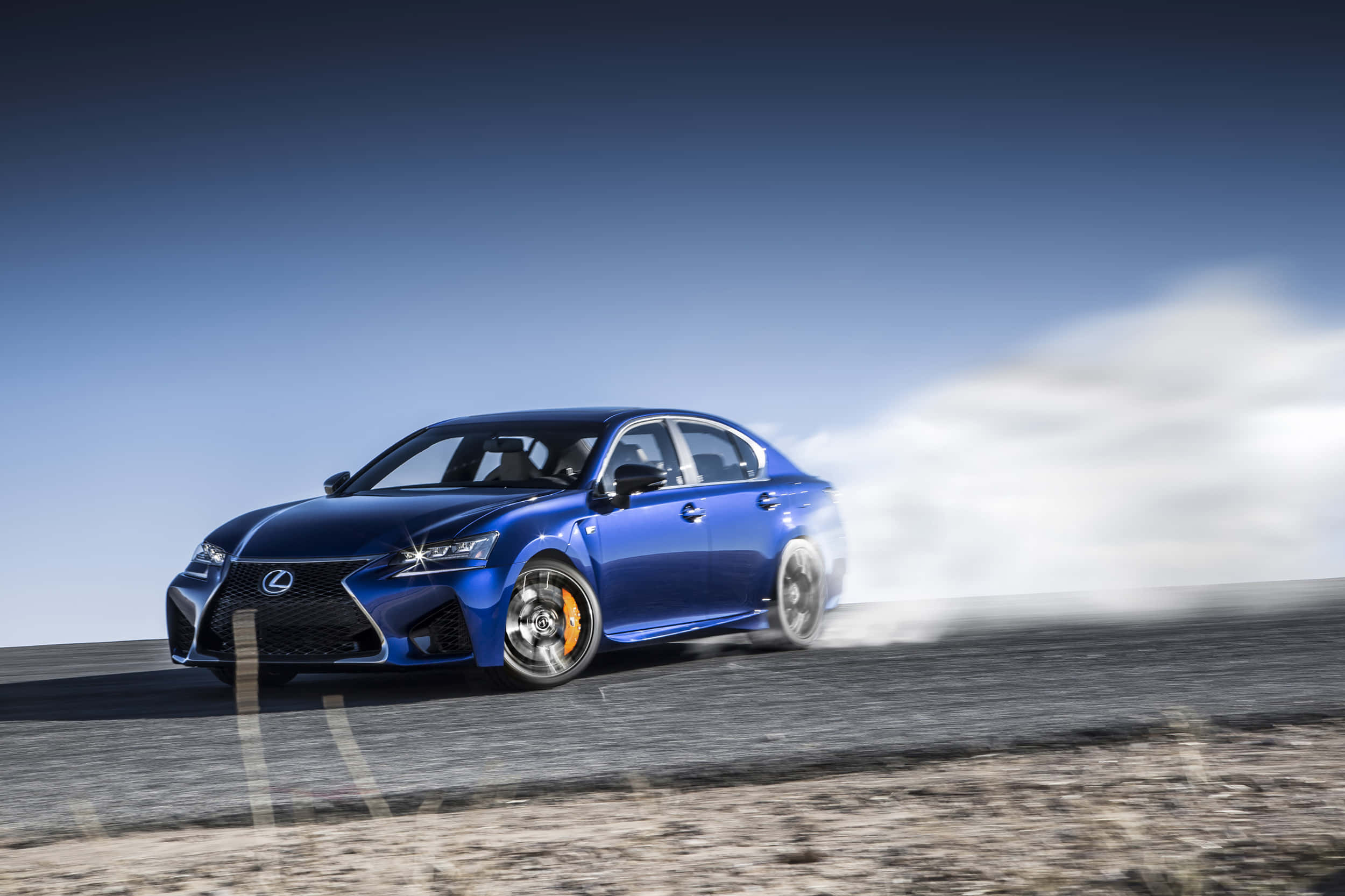 Lexus Gs F 桌布 壁紙 — 一輛充滿活力的藍色Lexus運動轎車在晴朗天空下的沙漠道路上甩尾，後輪冒出滾滾濃煙。